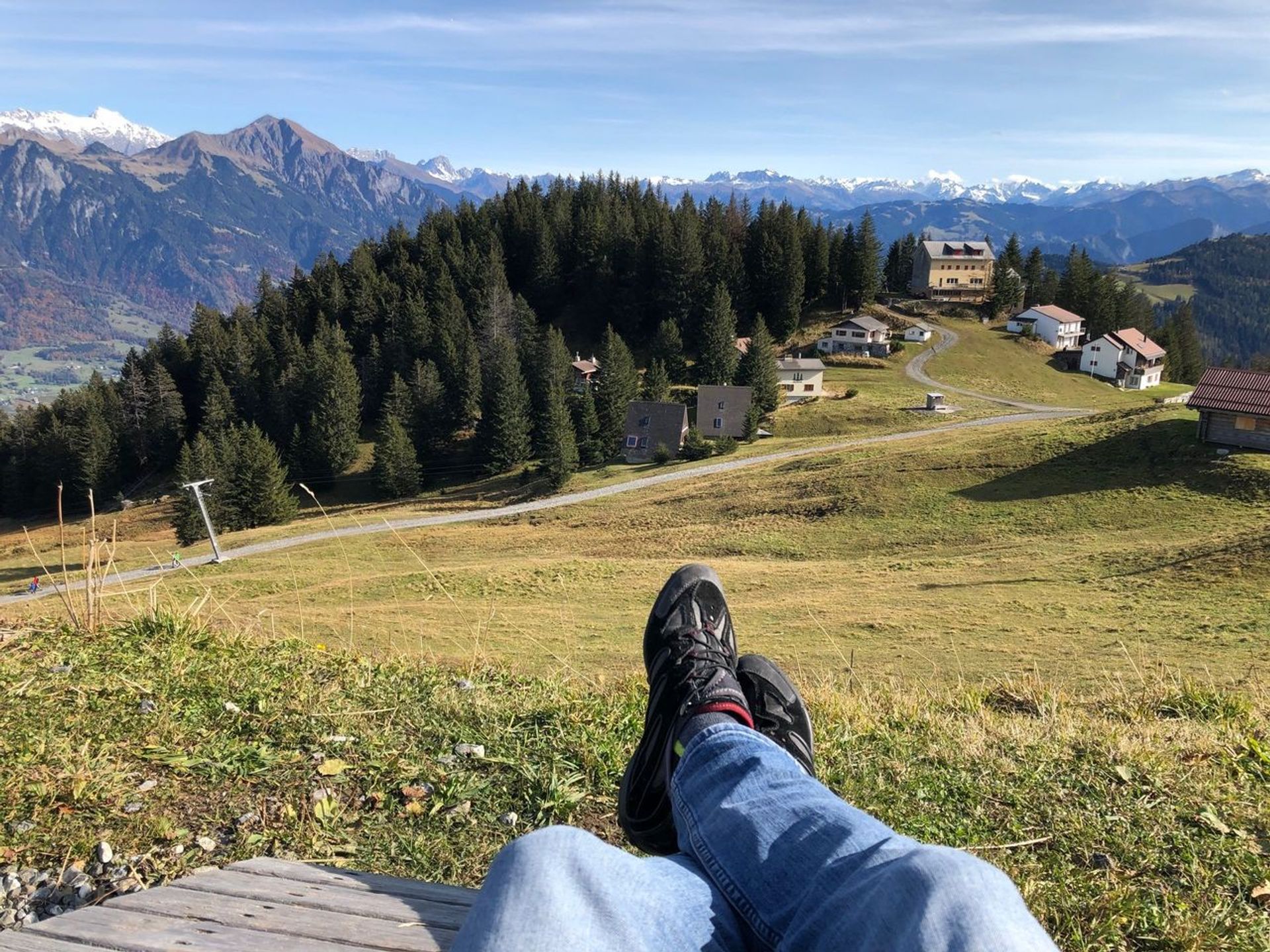 Im Vordergrund überkreuzte beine mit Wanderschuhen, dahinter gründe Berglandschaft mit vereinzelten Häusern