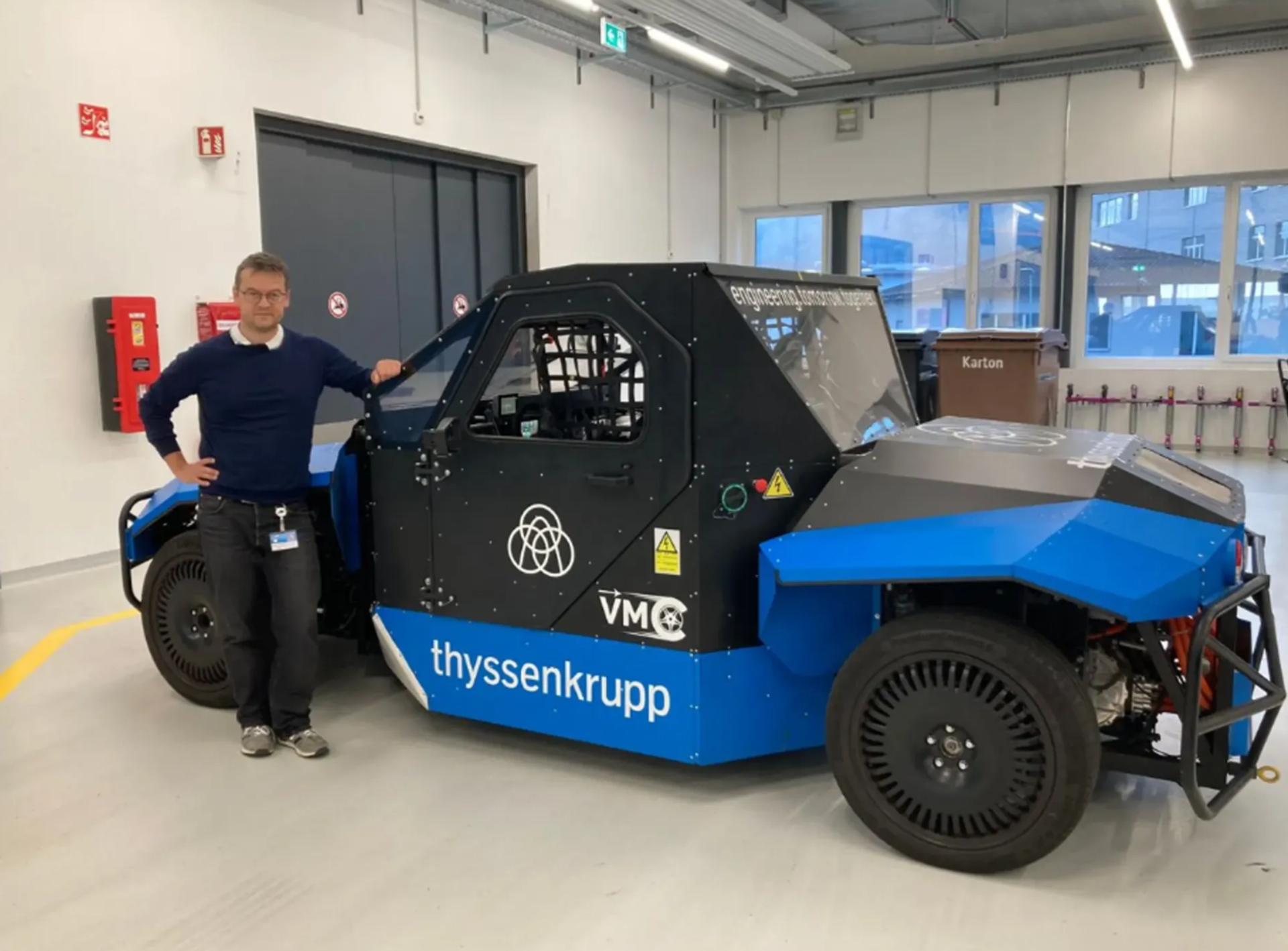 Male-presenting person stands in a workshop next to a blue-and-black vehicle prototype featuring the thyssenkrupp logo.