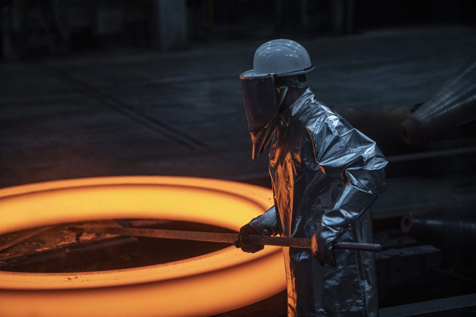 Ein Arbeiter in silberner Hitzeschutzkleidung und Schutzhelm steht in einer dunklen Industriehalle neben einem orange glühenden Metallring und hält eine lange Metallstange.