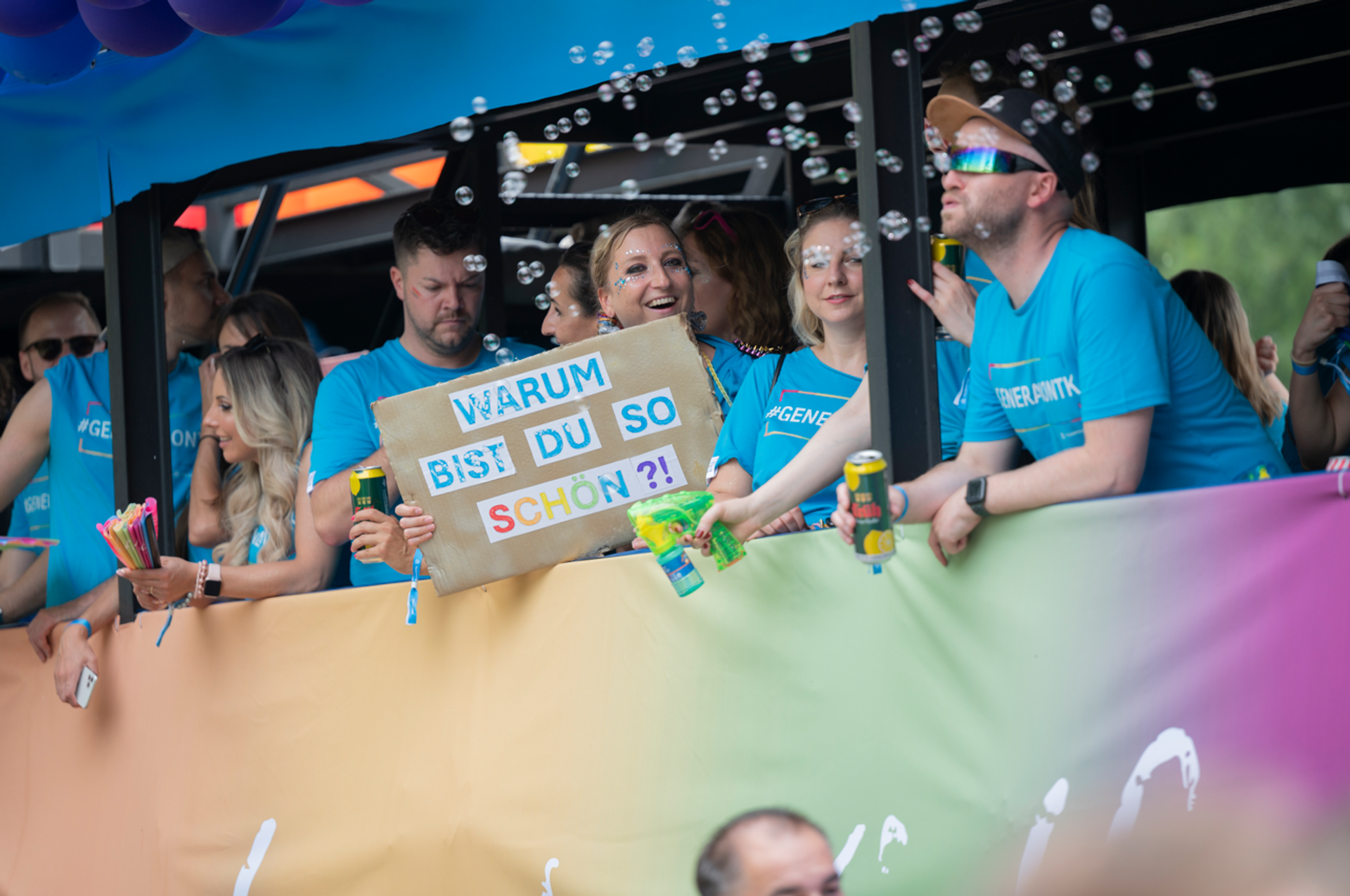 Menschen in blauen T‑Shirts stehen auf einem bunt geschmückten Pride‑Truck. Eine Person hält ein Schild mit der Aufschrift „Warum bist du so schön?!“. Über der Gruppe schweben Seifenblasen.