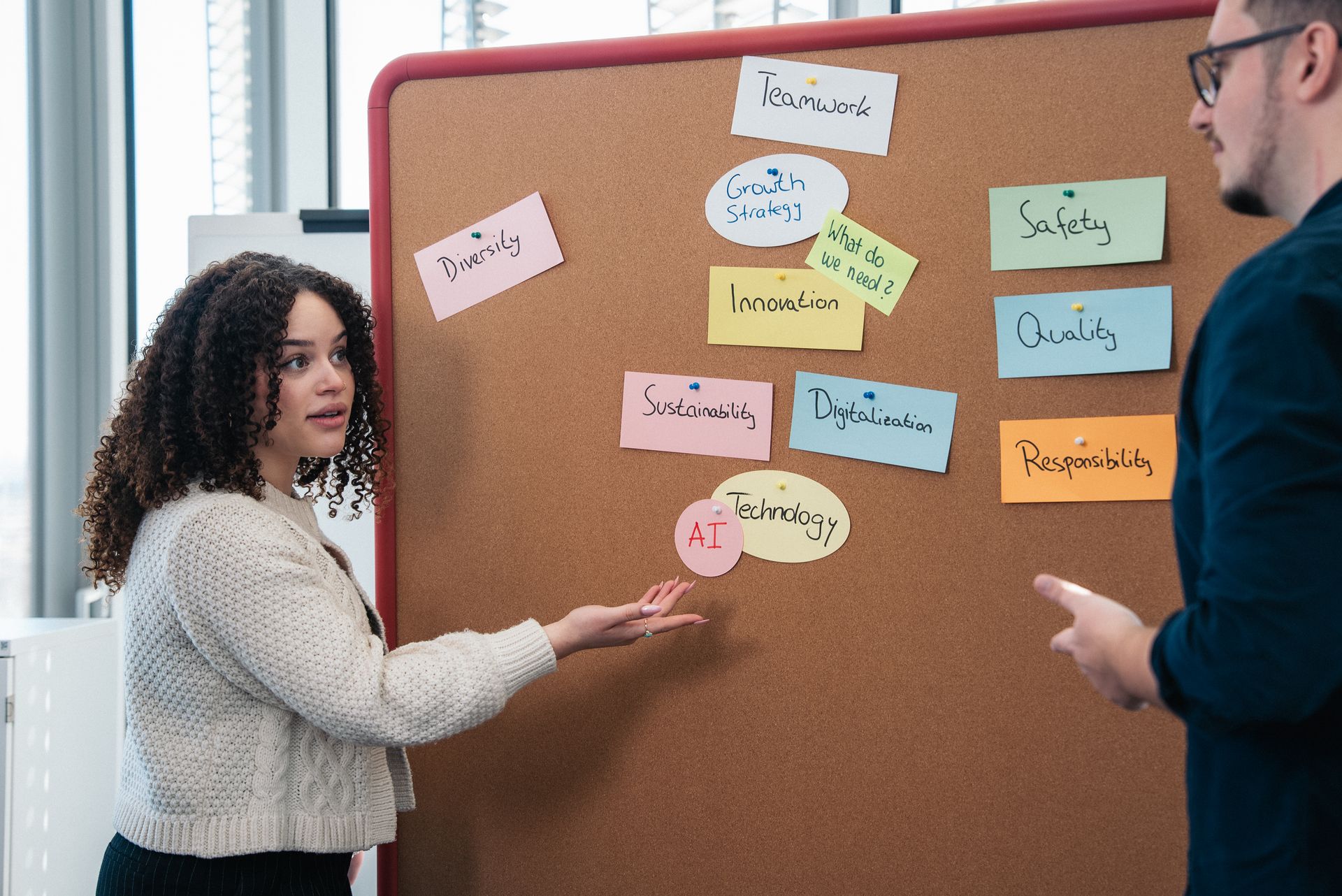 Eine junge Frau präsentiert Inhalte auf einer Pinnwand, die mit farbigen Kärtchen zu Themen wie Teamwork, Innovation, Technologie und Diversität bestückt ist. Neben ihr steht ein Kollege und hört aufmerksam zu.
