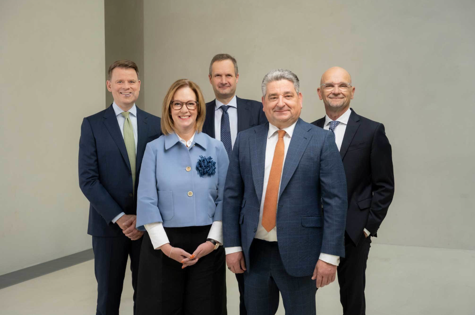 Fünf Mitglieder des Management-Teams stehen in Businesskleidung für ein professionelles Gruppenfoto in einem modernen Büro mit neutralem Hintergrund.