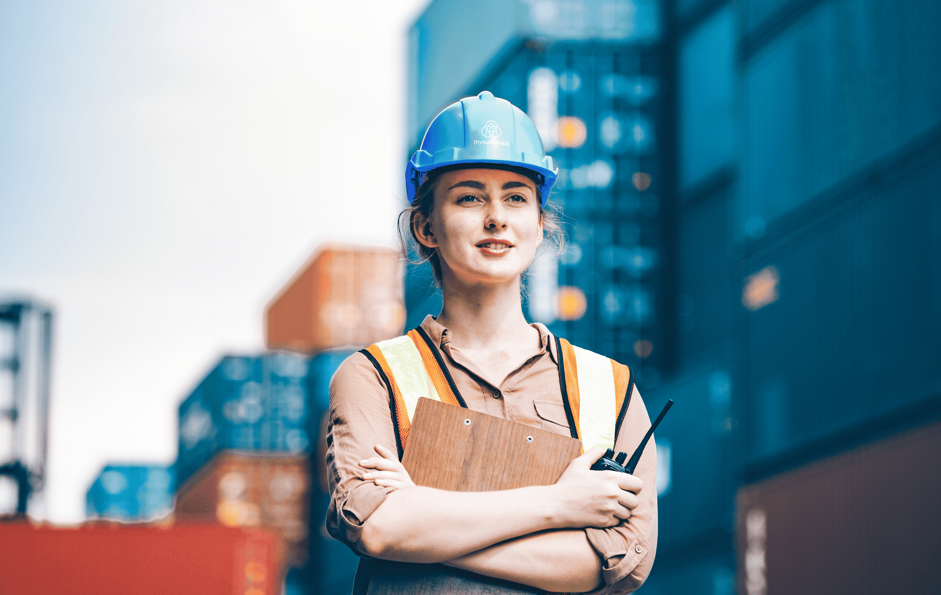 Junge Bauarbeiterin mit blauem Schutzhelm und Arbeitskleidung steht vor Stadtgebäuden.