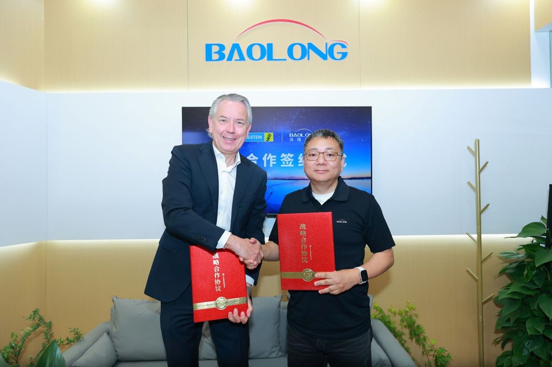 Two business-partner shaking hands in front of BALONG company press-wall, while holding red folders.