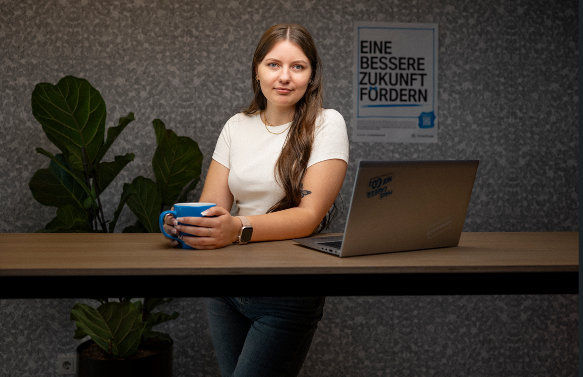 Frau sitzt an einem weißen Tisch mit geöffnetem Notizbuch, im Hintergrund Motivationsposter und goldener Ballon in Form einer Eins.