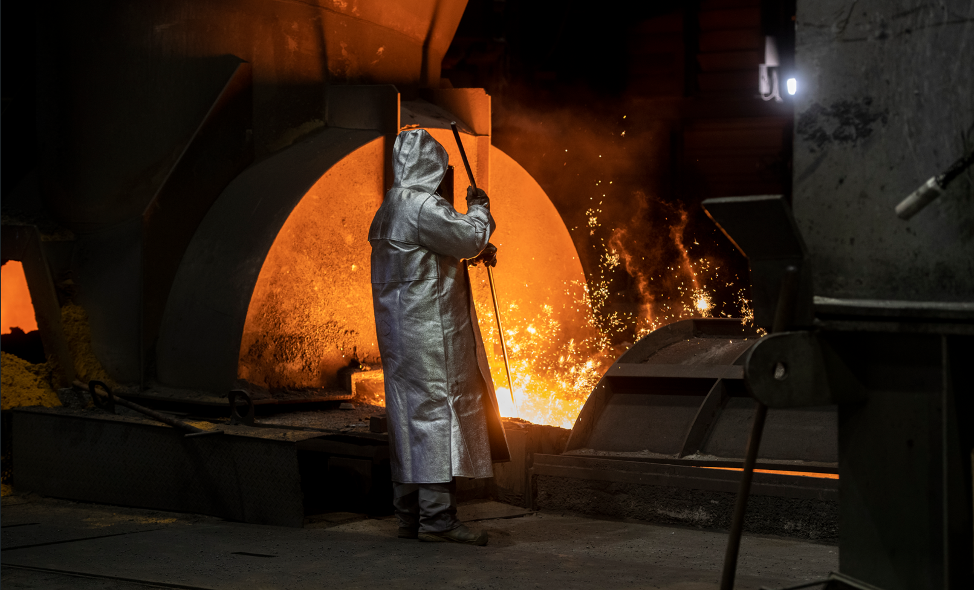 Arbeiter in Schutzkleidung arbeitet vor glühendem Schmelzofen.