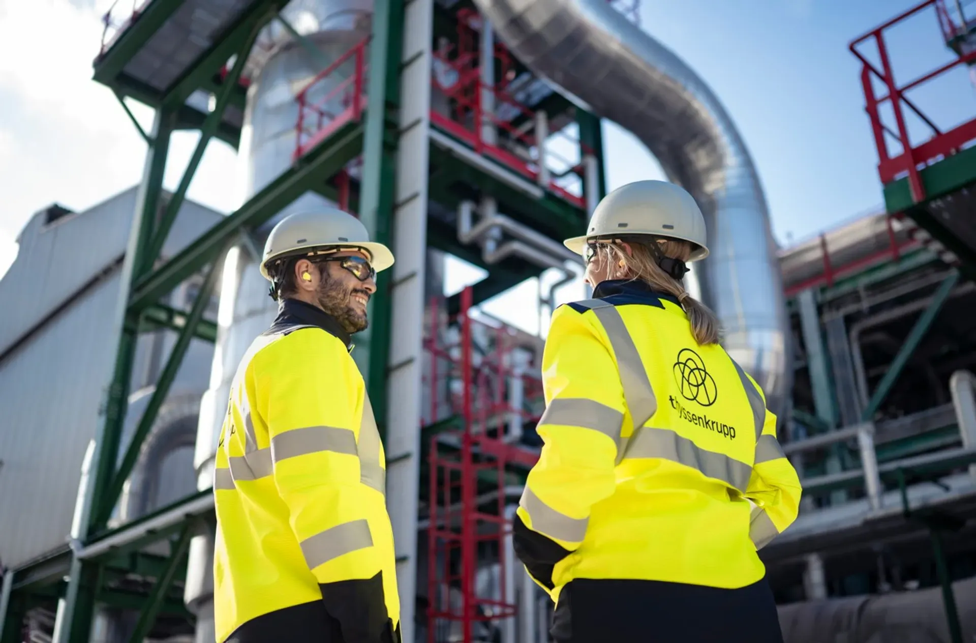 thyssenkrupp engineers in high-visibility jackets reviewing technical documents together on an industrial production floor