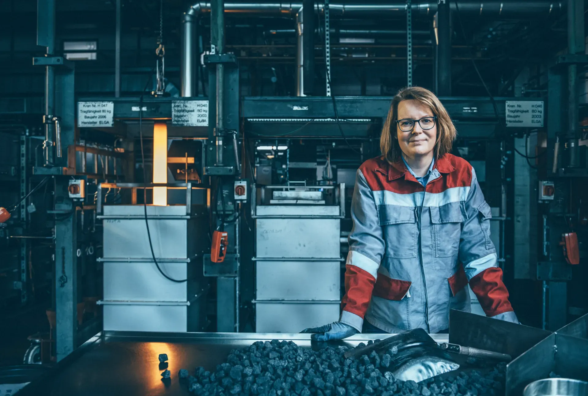A person wearing a grey and red work jacket stands at a table with black material pieces in an industrial production hall, with machinery and piping visible in the background.