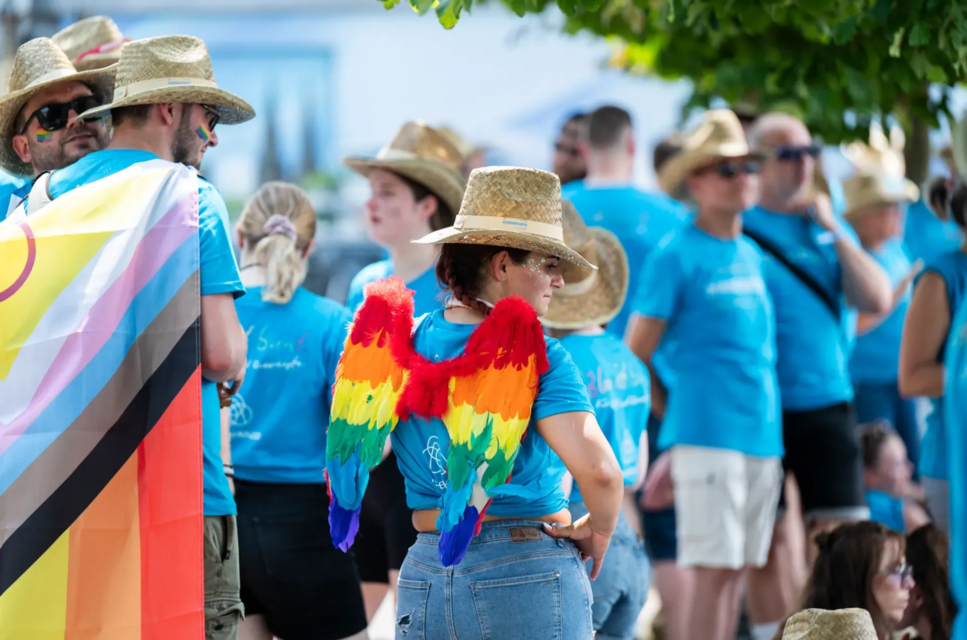 group of employees at a Pride parade wearing matching blue thyssenkrupp t-shirts and straw hats, one person wearing rainbow feather wings on their back, with a progress pride flag visible in the foreground