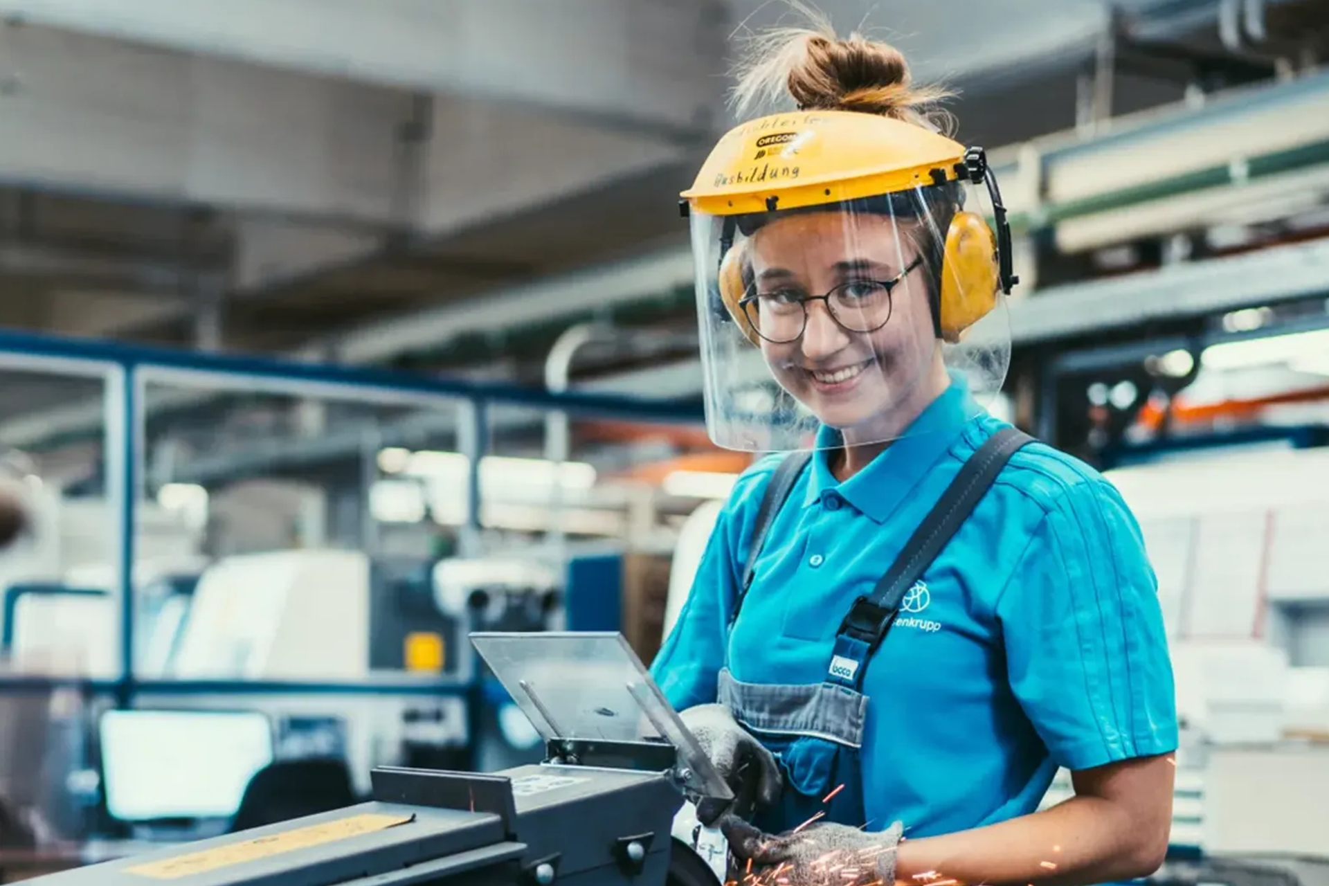 Eine Frau in Schutzkleidung arbeitet mit einer Maschine, die Funken erzeugt – vermutlich beim Schleifen oder Schneiden von Metall. Die Umgebung ist eine industrielle Werkstatt.