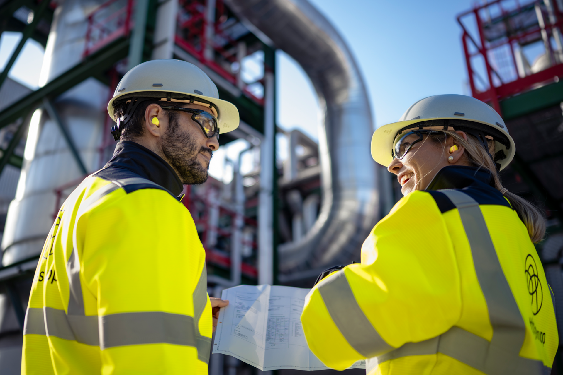 Zwei thyssenkrupp Beschäftigte in Warnschutzkleidung besprechen technische Unterlagen in einer Industrieanlage.