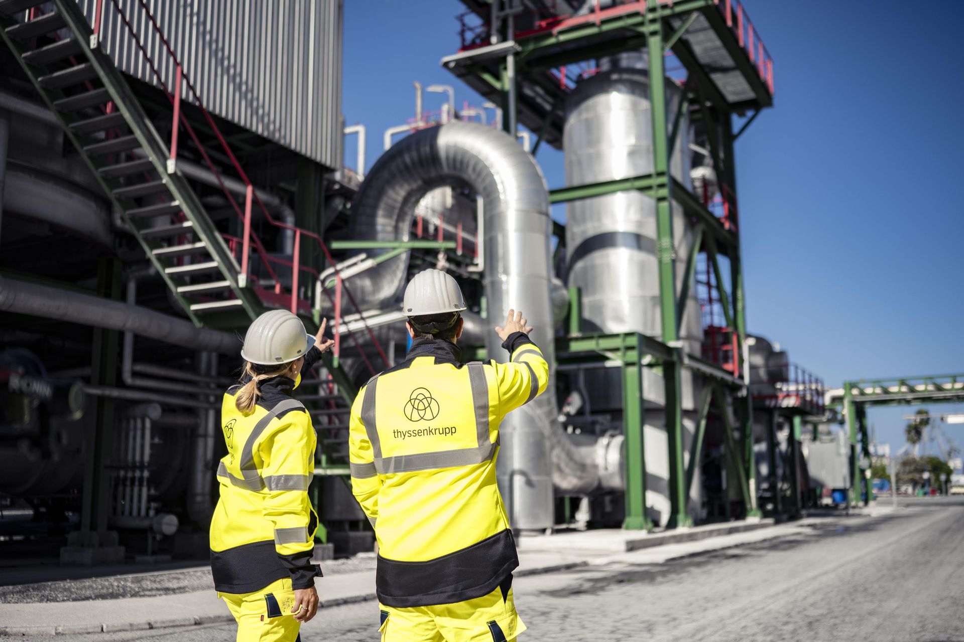 Zwei Mitarbeitende von thyssenkrupp mit Schutzhelmen und Warnkleidung vor einer industriellen Chemieanlage.