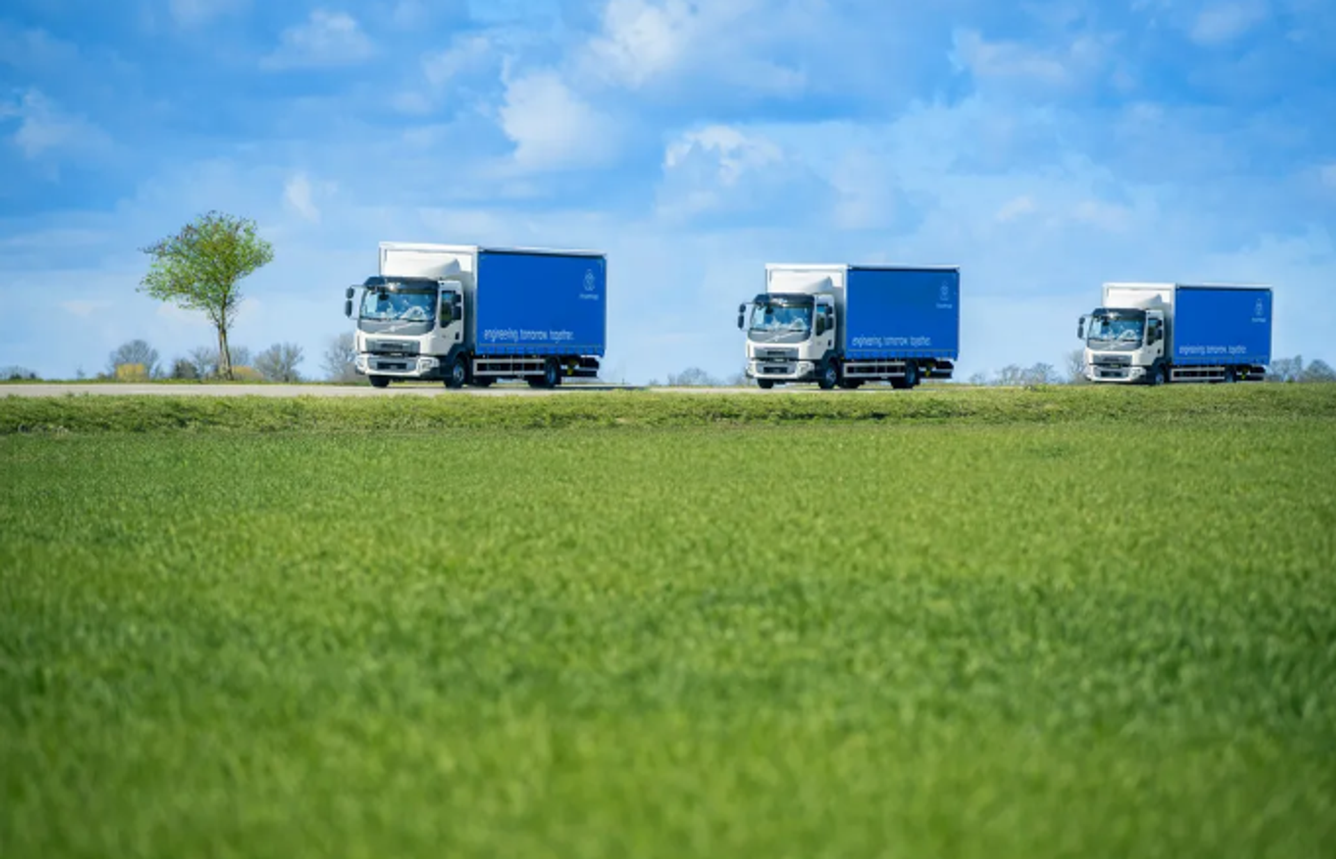 Three blue trucks drive one behind the other along a road through a green landscape under a blue sky.