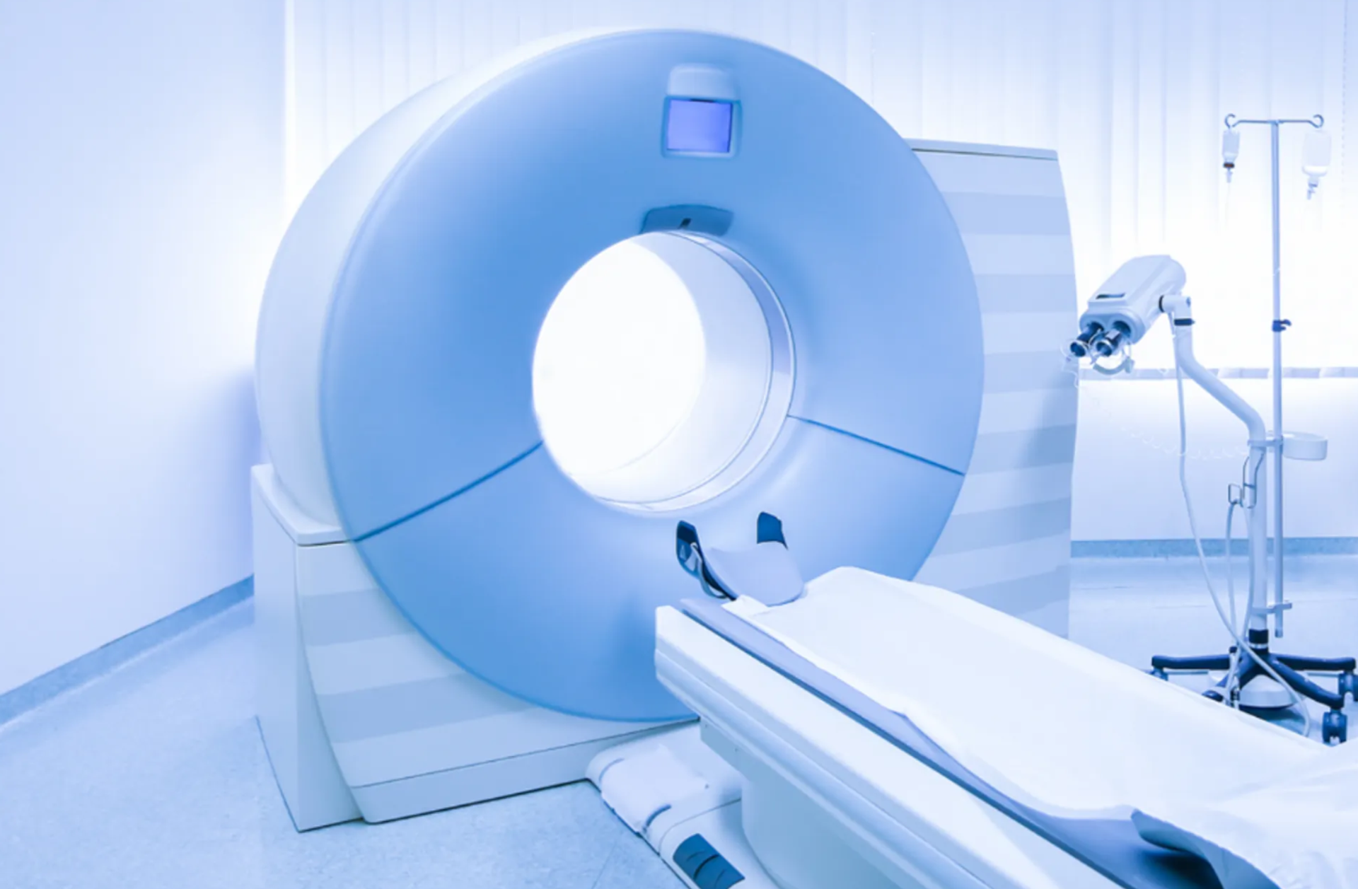CT scanner in a bright examination room with a patient table, IV stand, and medical equipment.