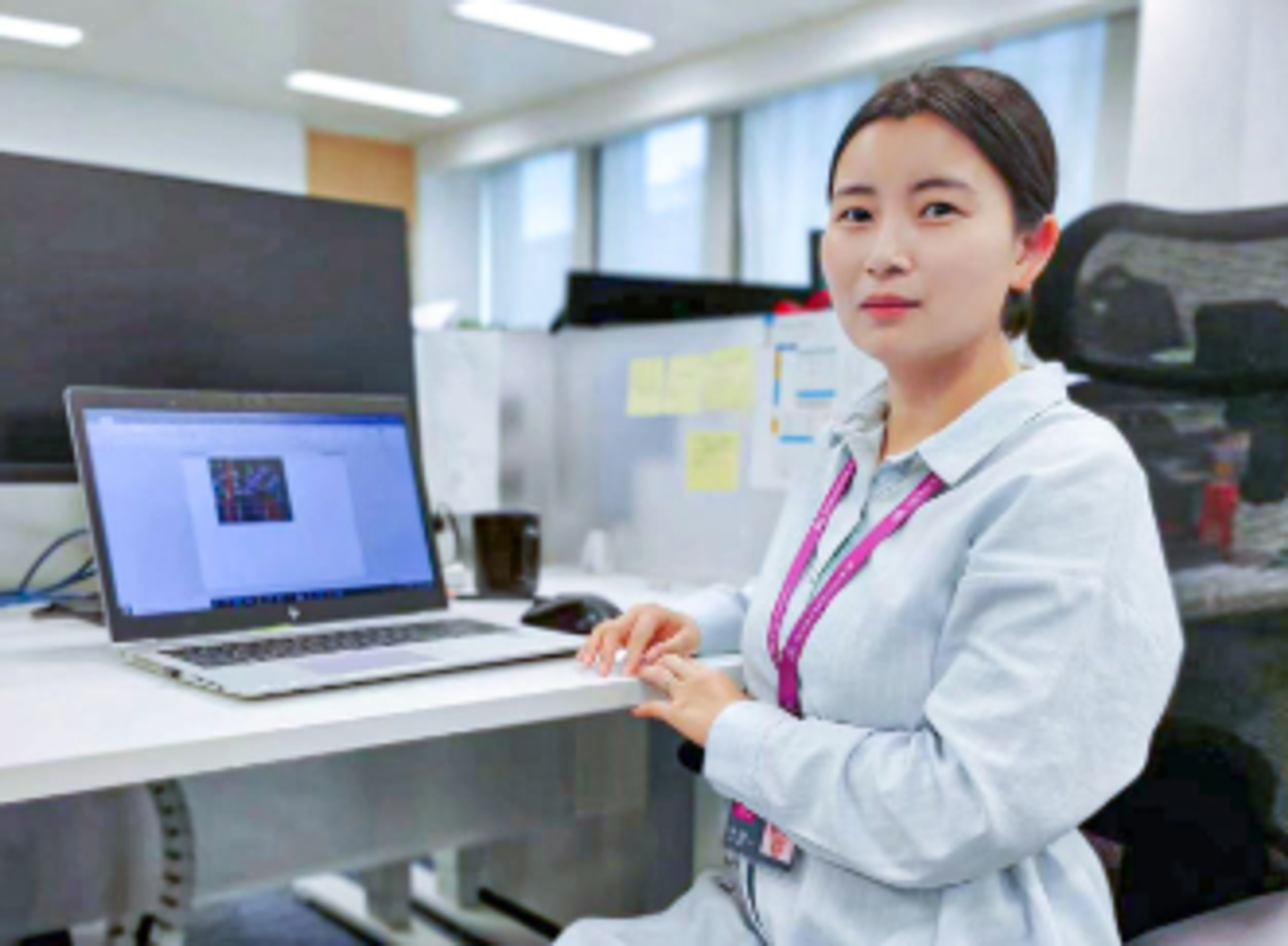 Yuan-Yan Shao: Frau mit braunen Haaren, zu einem Zopf gebunden, sitzt in weißen langem Oberteil auf einem Schreibtischstuhl vor ihrem Laptop