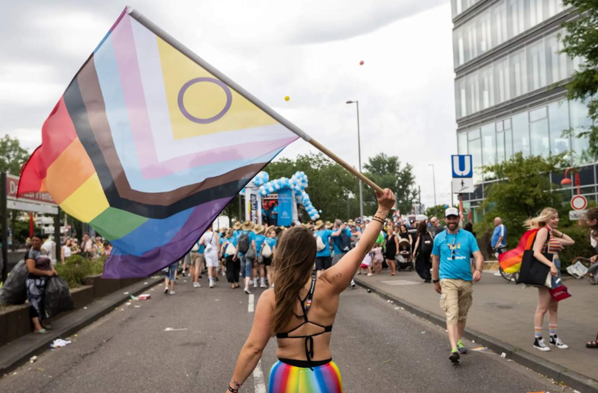 A person at a CSD parade waves a large Progress Pride flag. In the background, a crowd moves along a city street.