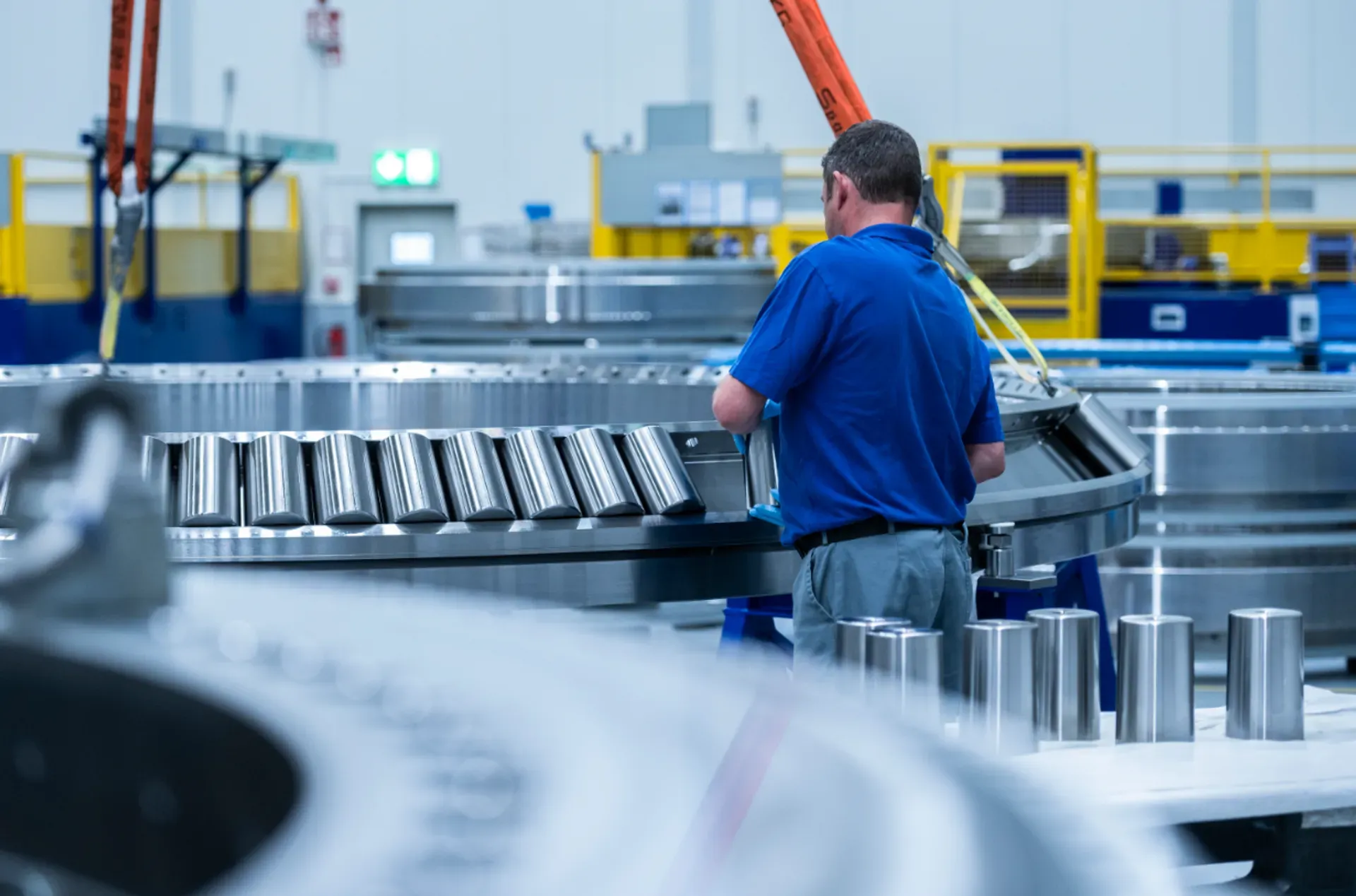 Ein thyssenkrupp Mitarbeiter arbeitet in einer Produktionshalle an einem großen Wälzlager mit sichtbaren Rollen; weitere Metallbauteile und Fertigungsanlagen befinden sich im Hintergrund.