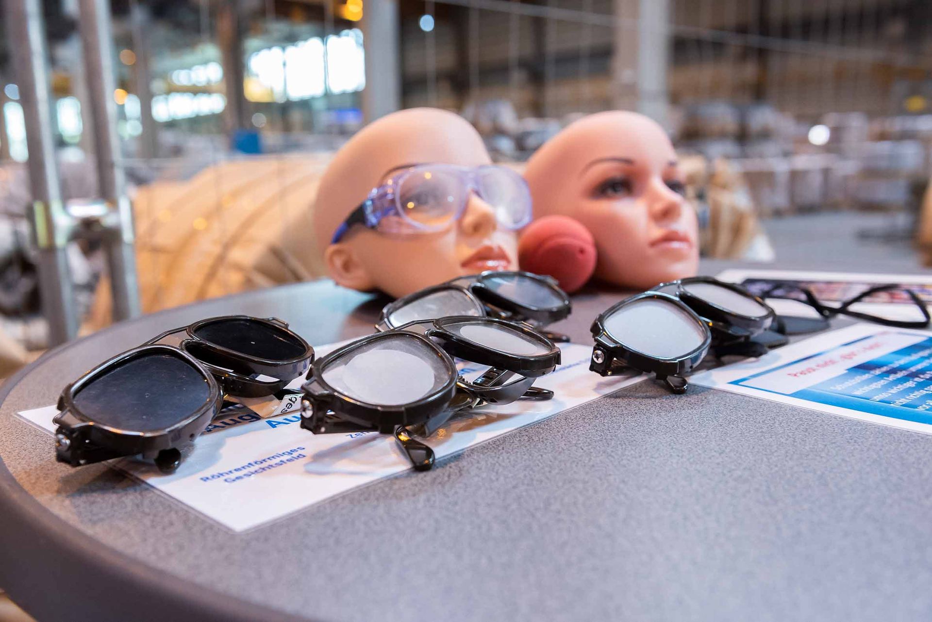 Simulationsbrillen und zwei Übungsköpfe mit Schutzbrille auf einem Tisch bei einer Arbeitsschutzaktion in einer Werkshalle.