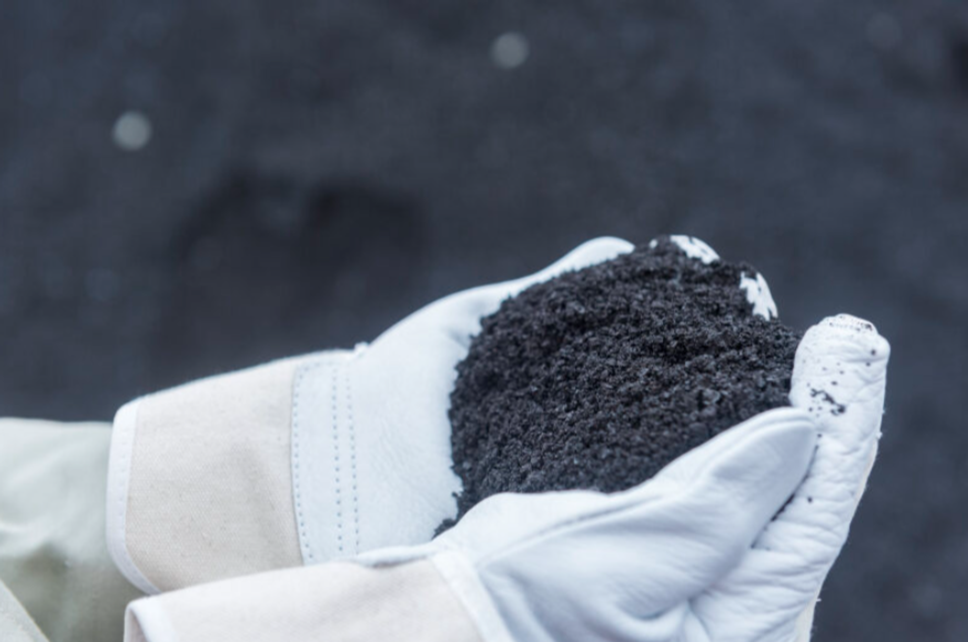 Hand mit Schutzhandschuh hält dunkles Recyclingmaterial oder Schlacke aus der Stahlproduktion als Rohstoff für die industrielle Wiederverwertung.