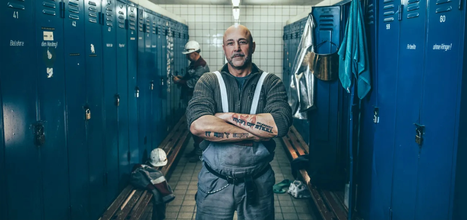 Industriearbeiter in Umkleidekabine mit blauen Spinden, tätowiertem Schriftzug ‚Man of Steel‘ auf dem Arm 