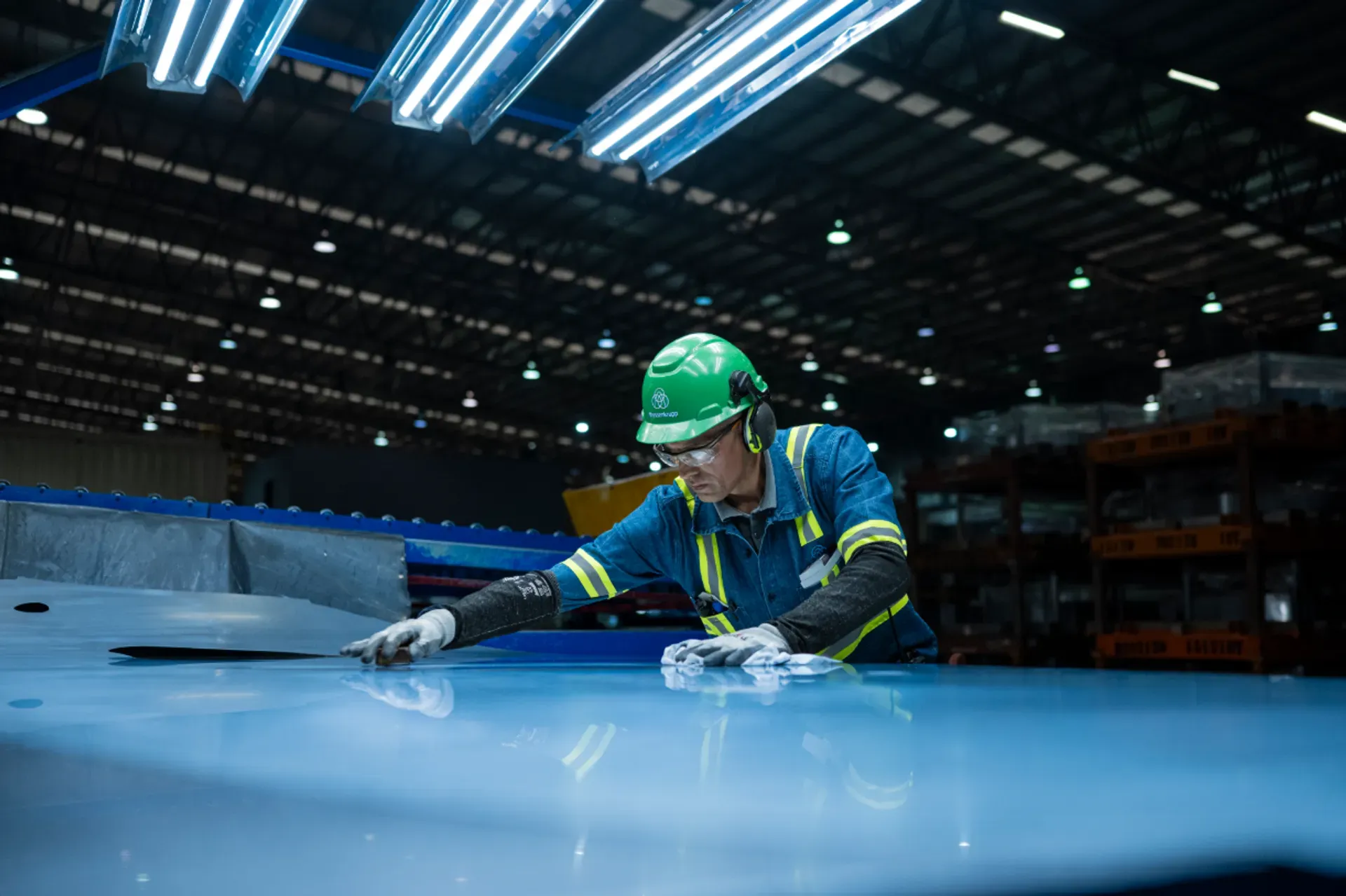 Eine Fachkraft mit Schutzhelm und Warnkleidung prüft die Oberfläche einer großen Metallplatte in einer Industriehalle.