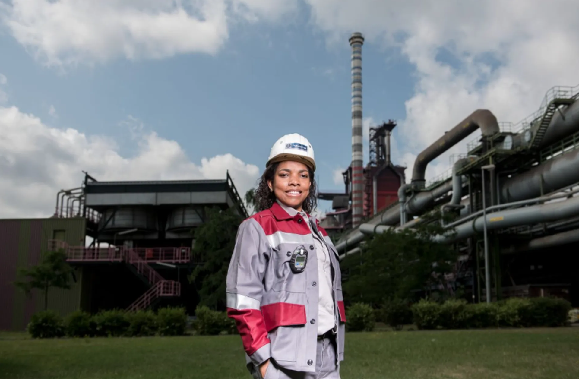 Weiblich gelesene Person in Arbeitskleidung und Schutzhelm steht vor einer industriellen Anlage mit Rohrleitungen und Schornstein.