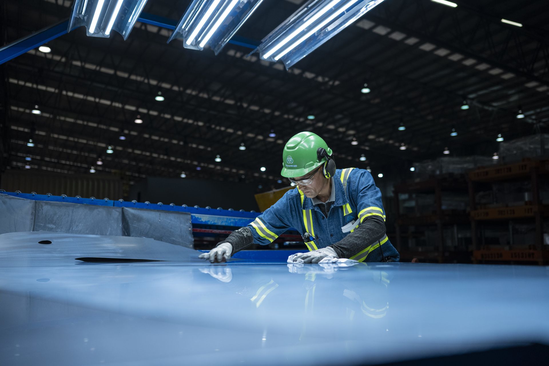 Mitarbeiter mit grünem Schutzhelm prüft eine große Metallplatte in einer Lagerhalle.