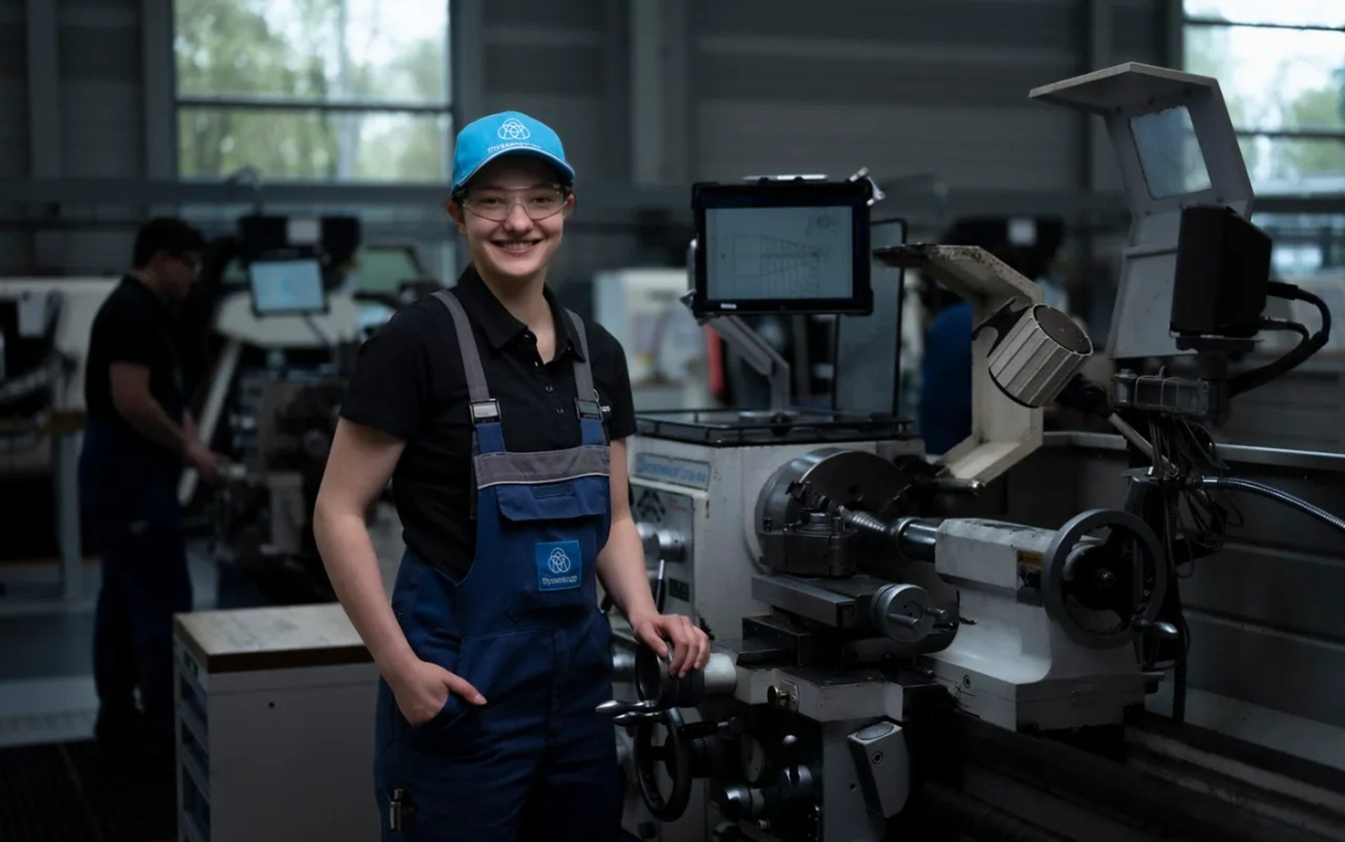 Junge Frau in blauer Arbeitskleidung und Basecap steht neben einer Drehmaschine in einer modernen Werkstatt.