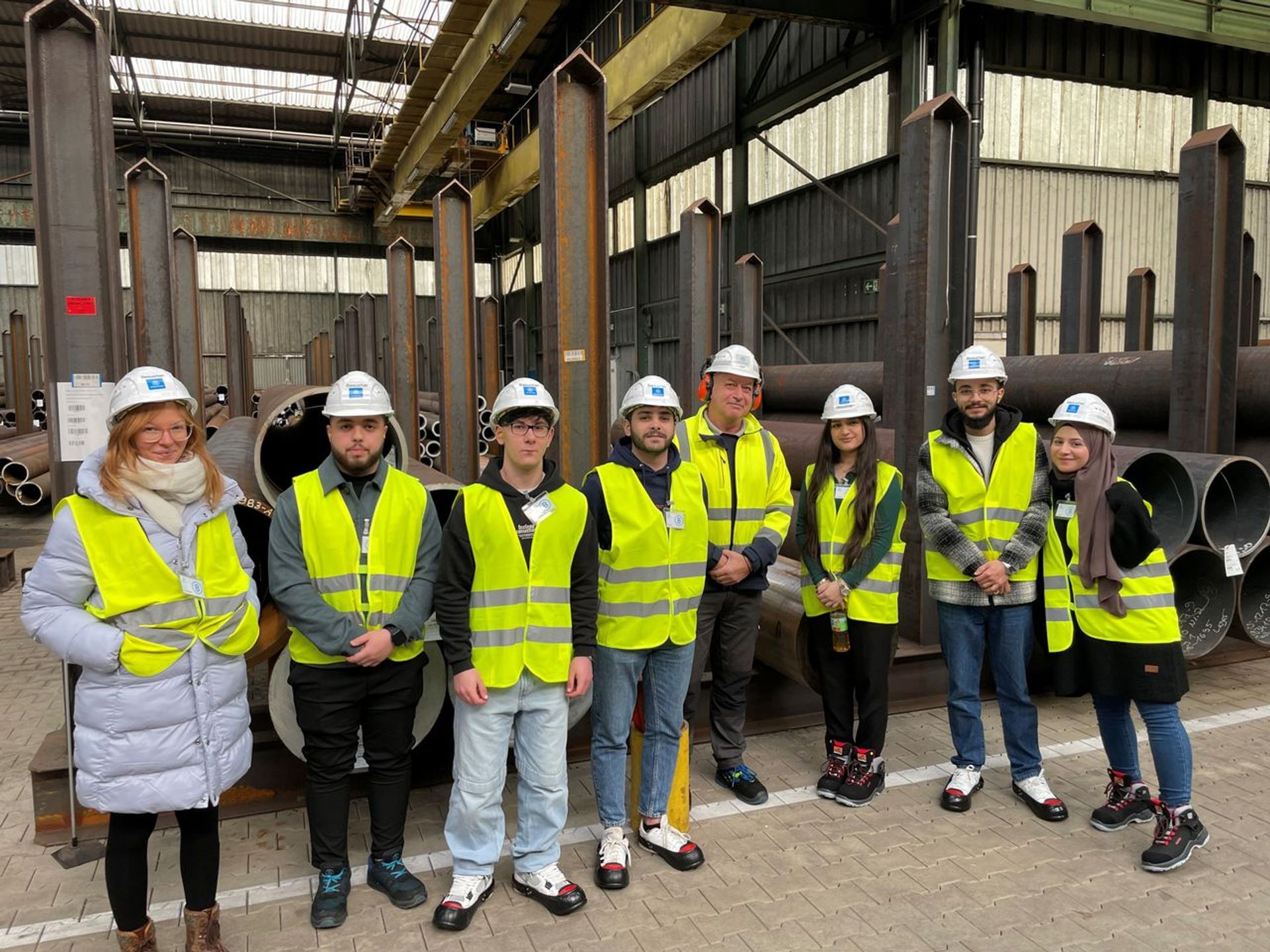 Eine Gruppe junger Menschen mit Schutzhelmen und Warnwesten steht in einer Industriehalle während einer Werksbesichtigung bei thyssenkrupp
