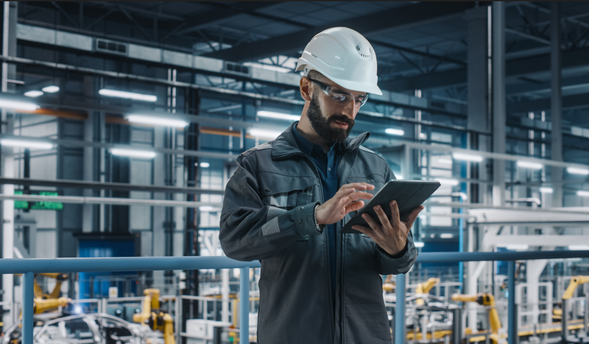 Eine Person mit Schutzhelm und Arbeitsjacke steht in einer modernen Industriehalle und bedient ein Tablet. Im Hintergrund sind Maschinen, Förderanlagen und Metallkonstruktionen einer Produktionsumgebung zu sehen.