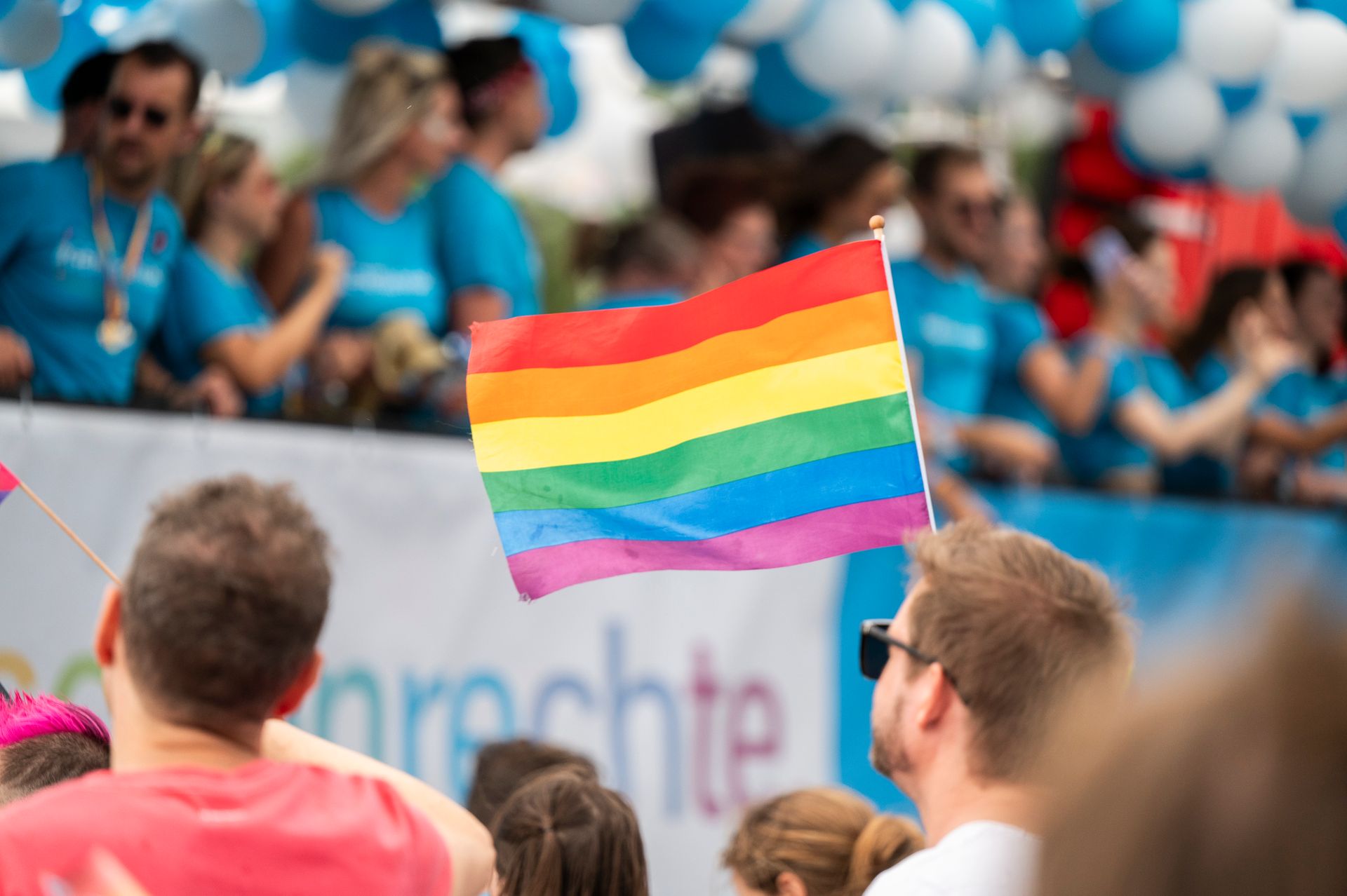 A rainbow Pride flag is held up in a lively parade setting, surrounded by people in colorful clothing and blue‑and‑white balloons.
