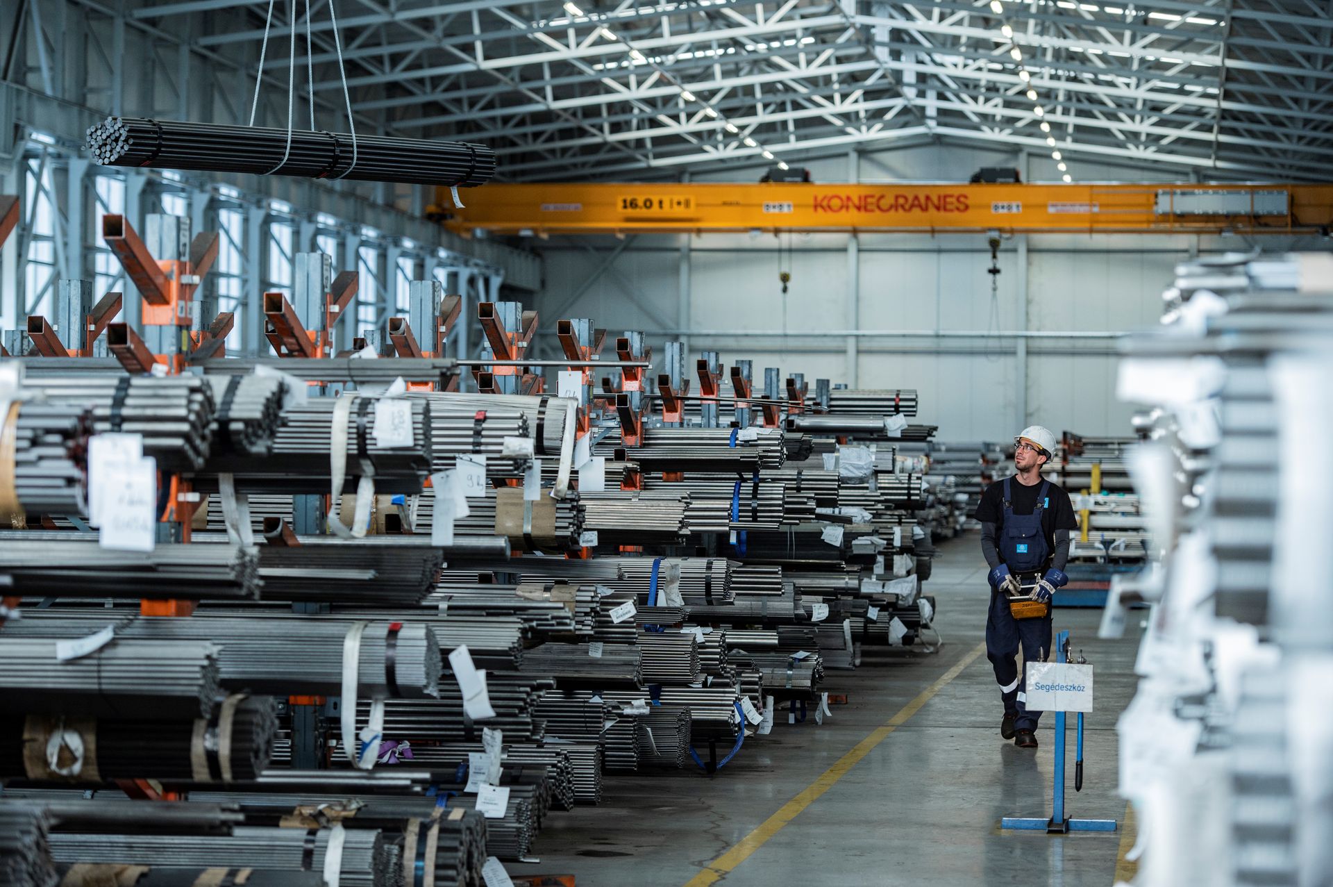 Mehrere Metallstangen sind in einer Lagerhalle und ein paar Metallstangen werden von einem Mitarbeiter mit Elektronik transportiert.