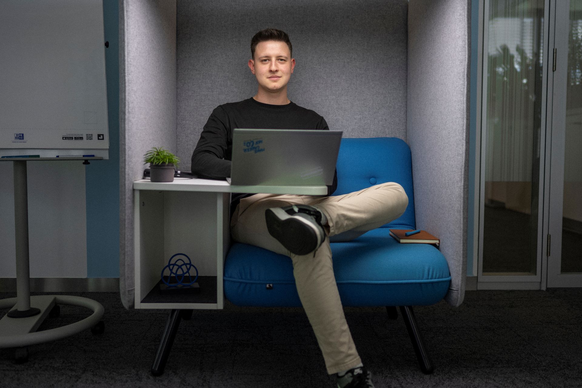 Person sitzt in einer modernen Bürobox auf einem blauen Sessel und arbeitet am Laptop.