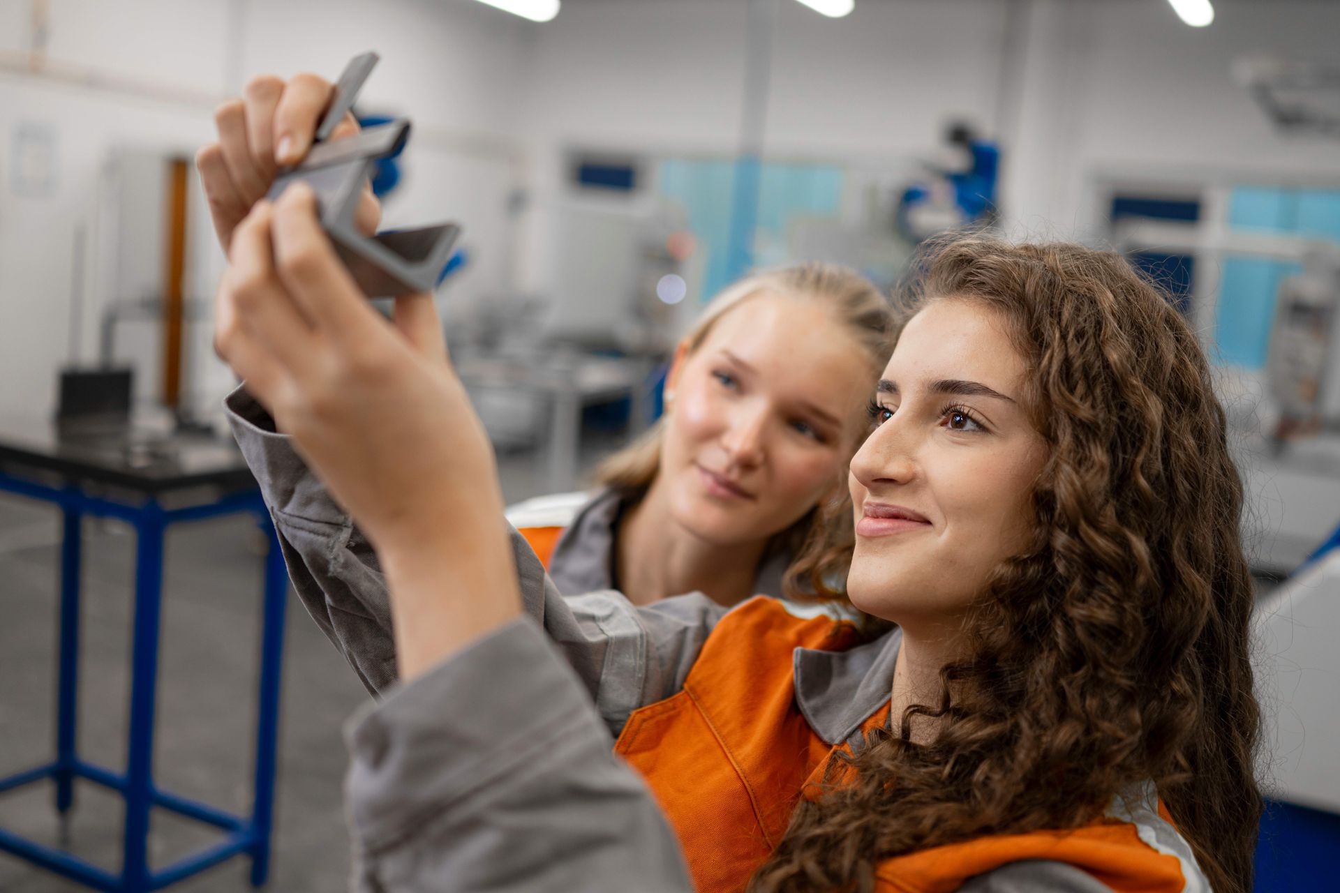 Zwei junge Auszubildende stehen in einer Werkstatt und betrachten gemeinsam ein Metallteil, das eine von ihnen in den Händen hält. Beide wirken konzentriert und interessiert, während sie in ihrer Arbeitskleidung eine praktische Aufgabe lösen.
