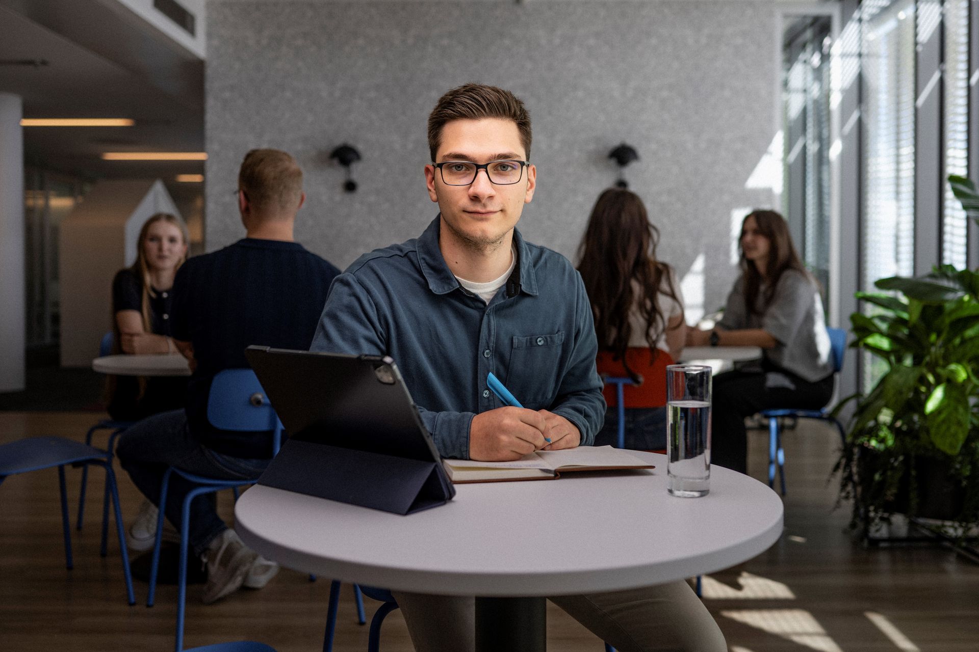 Junger Mann schreibt in ein Notizbuch an einem Tisch mit Tablet und Wasser, im Hintergrund offene Bürofläche mit mehreren Personen.