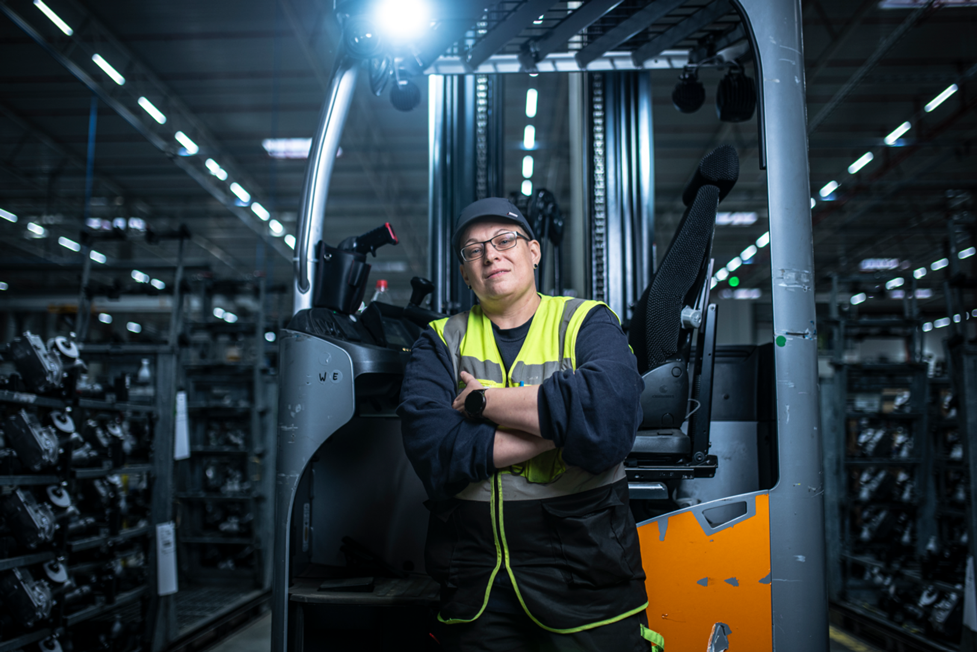 Person in Sicherheitsweste steht mit verschränkten Armen vor einem Gabelstapler in einer großen Industriehalle. Im Hintergrund sind Regale mit Metallteilen zu sehen, die Halle ist gut beleuchtet mit langen LED-Lichtstreifen an der Decke.
