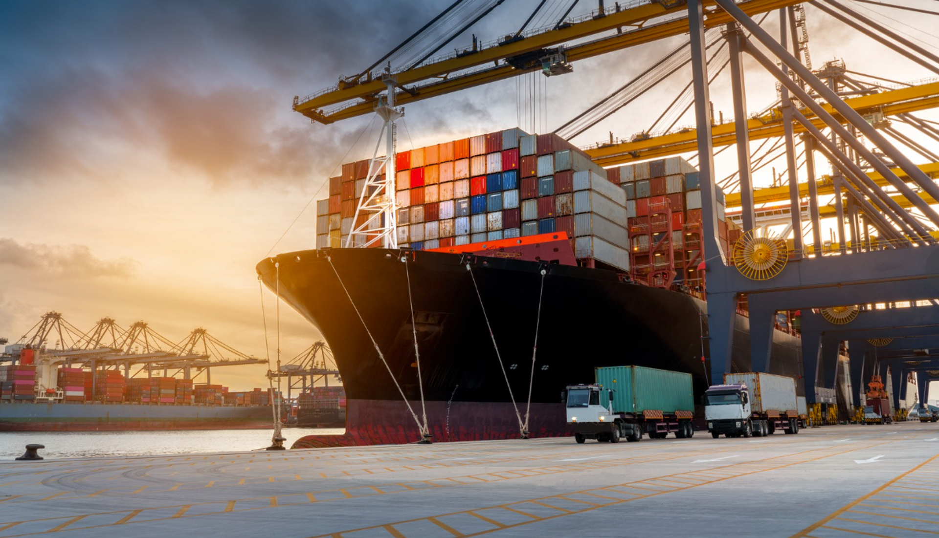 Nahaufnahme eines großen Containerschiffs an einem Umschlagshafen. Das Schiff liegt am Kai unter mehreren hohen Containerkränen, während farbige Container gestapelt und verladen werden.