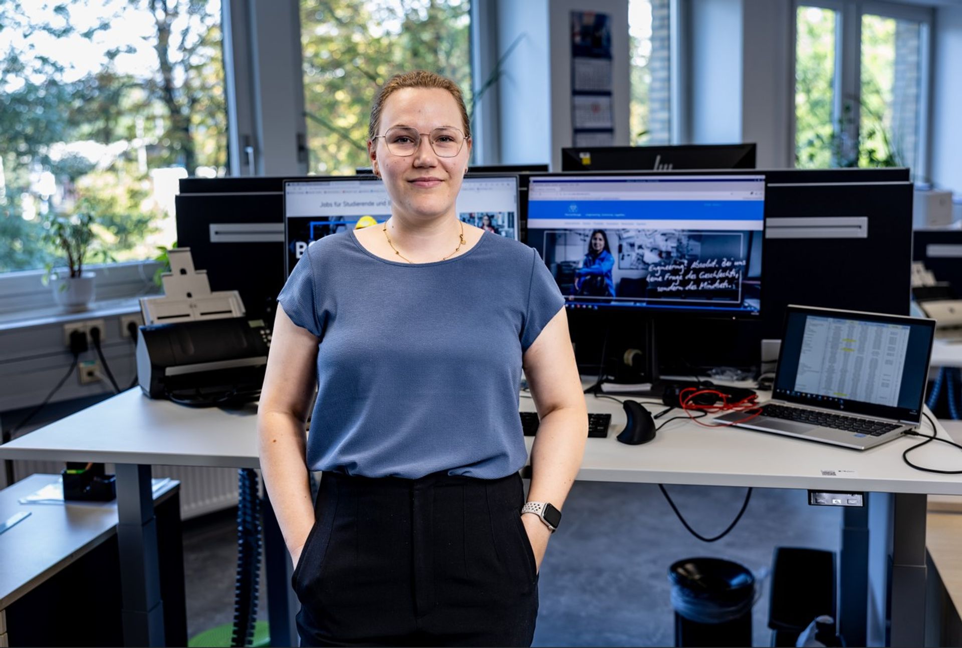 Mitarbeiterin steht in einem modernen Büro an einem höhenverstellbaren Schreibtisch mit Monitoren und Headset, im Hintergrund hängen Fotos und Poster an der Wand.