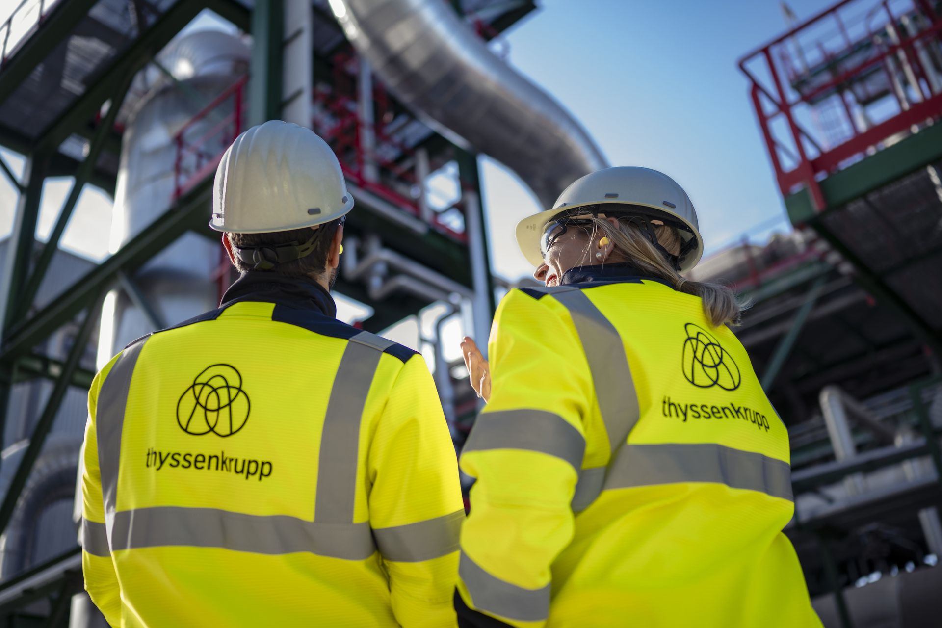 Zwei Mitarbeitende von thyssenkrupp in Schutzkleidung und Helmen vor einer Industrieanlage. Sie besprechen Arbeiten im Chemieanlagenbau vor Ort.