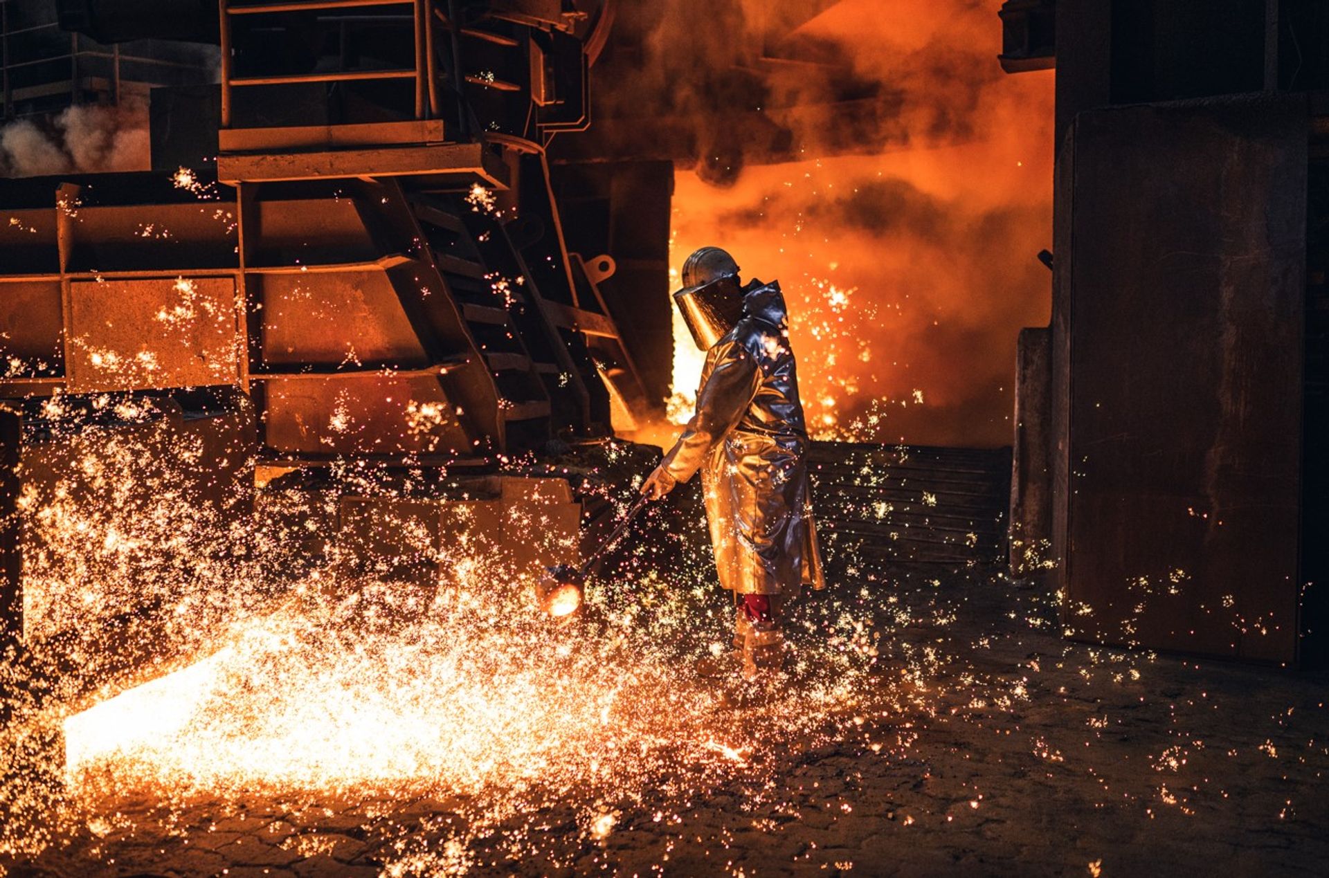 Industriearbeiter in Schutzkleidung steht in einer Stahlgießerei neben einem Strom aus glühend heißem Metall. Funken sprühen über den Boden, während im Hintergrund orangefarbene Flammen und Rauch die Szene erhellen.