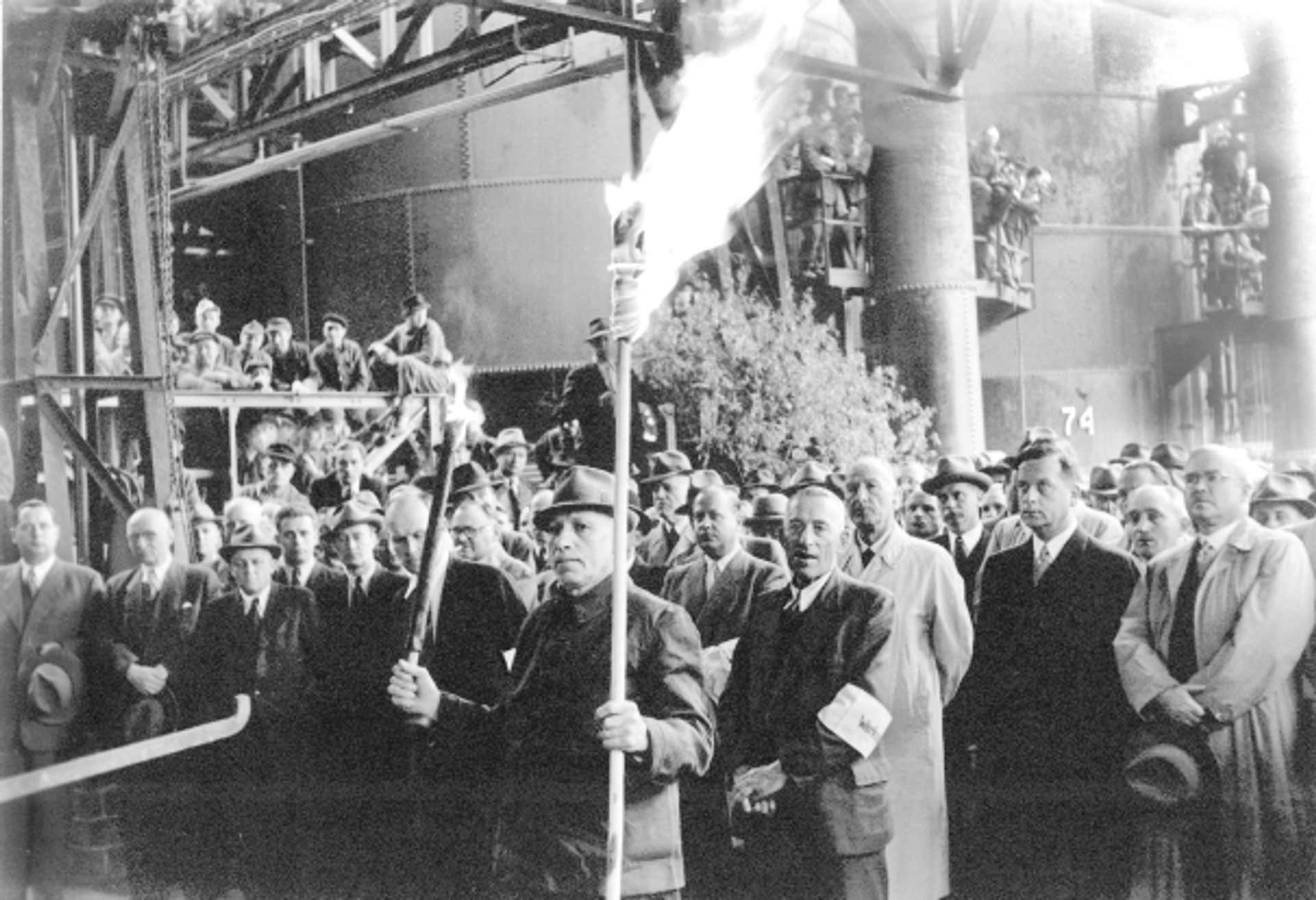 Feierstunde auf der Thyssen-Hütte am 7. Mai 1951, ein Arbeiter hält eine brennende Fackel vor versammelter Belegschaft.