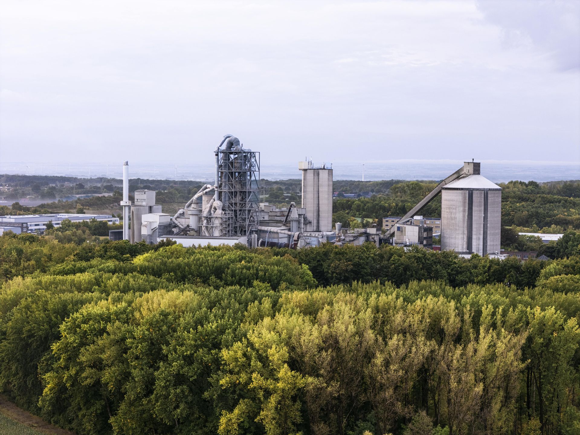 Zementwerk mit Industrieanlagen und Silos, umgeben von Bäumen und einem See im Vordergrund, als Beispiel für Technologien zur CO₂-Reduktion in der Zementproduktion.