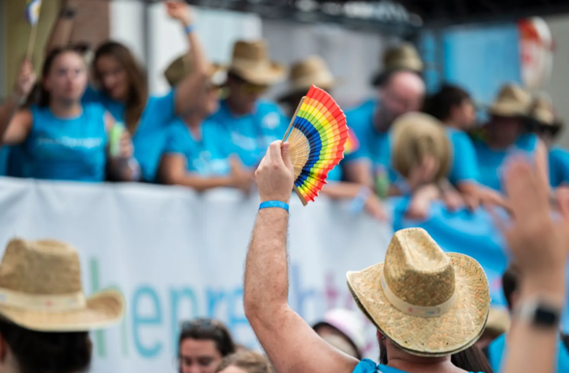A person wearing a straw hat holds up a rainbow fan at a Pride event. Other participants in blue T-shirts are standing in the background.