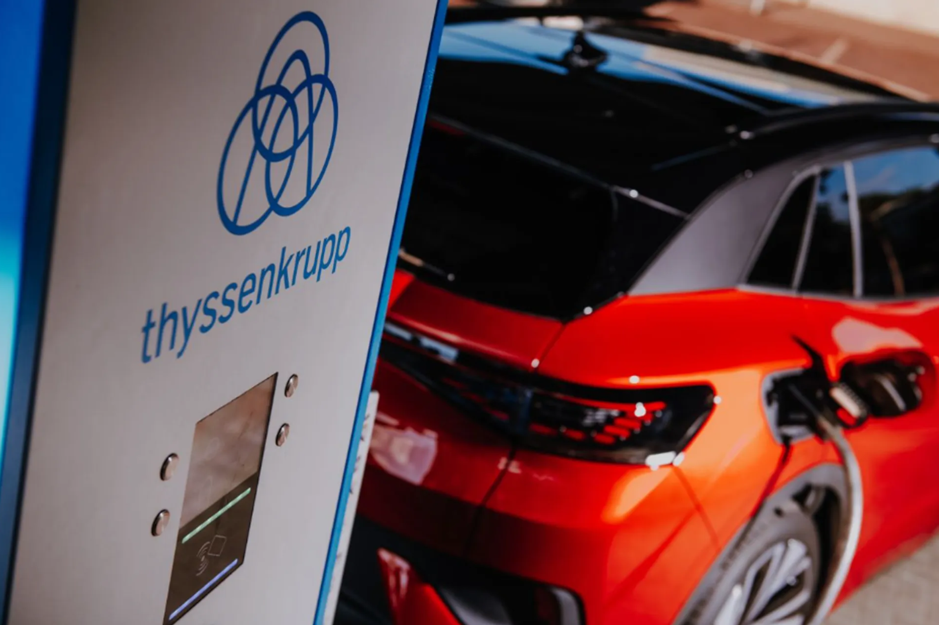 Ein rotes Elektrofahrzeug lädt an einer Ladesäule mit thyssenkrupp-Logo; im Vordergrund ist die Ladesäule, im Hintergrund das angeschlossene Fahrzeug zu sehen.