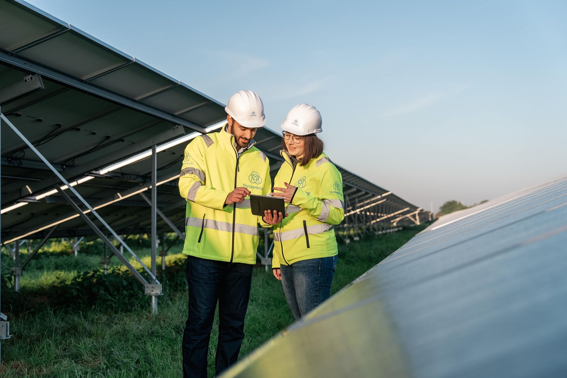 Zwei Mitarbeitende von thyssenkrupp mit Schutzhelmen und Warnkleidung stehen in einem Solarpark und betrachten gemeinsam ein Tablet.