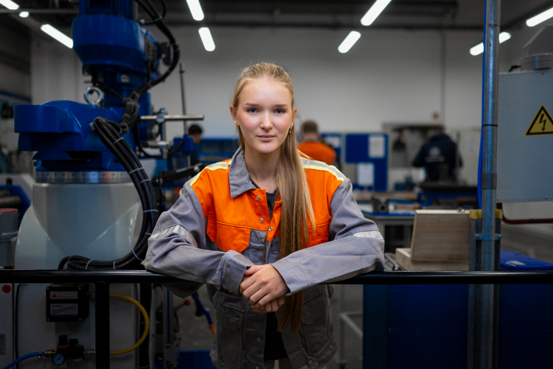 Frau steht in einer großen Produktionshalle neben blauen Metallgestellen, trägt dunkle Kleidung und Warnweste.