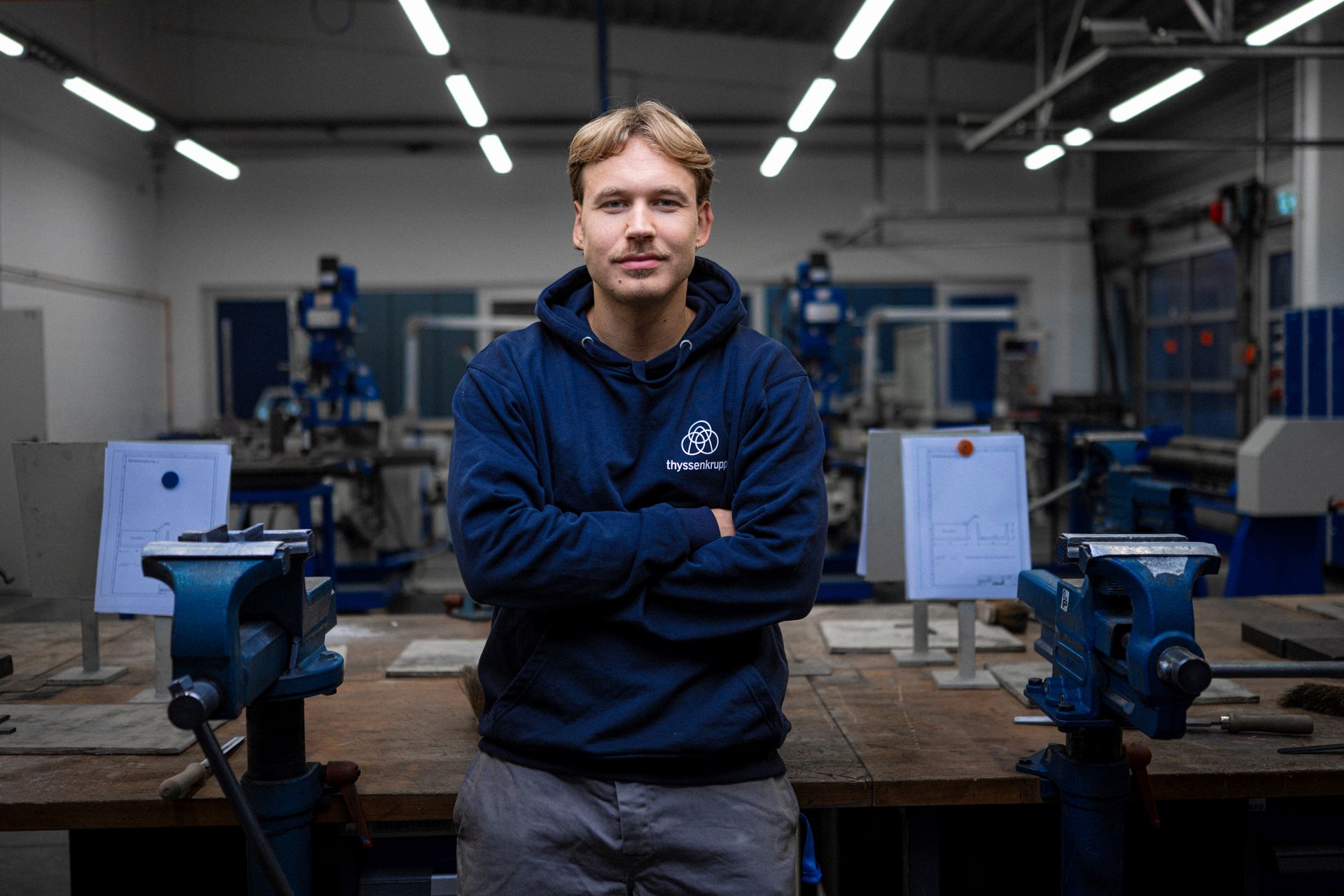 An apprentice stands in a workshop between vices and machines and looks towards the camera.