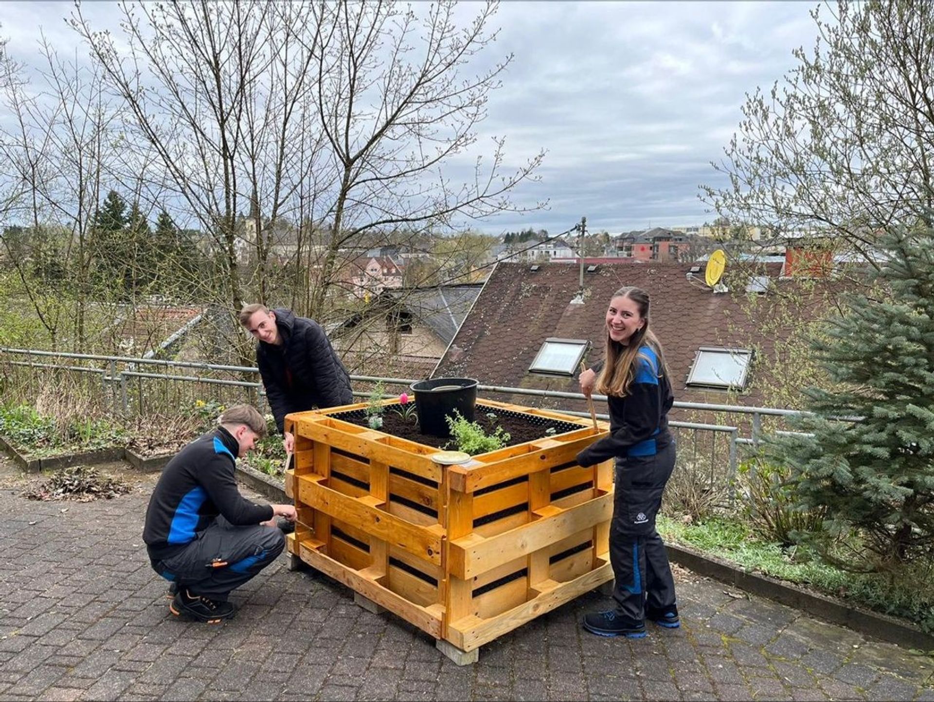 Drei Auszubildende von thyssenkrupp bauen gemeinsam ein Hochbeet aus Holz auf einer Terrasse.