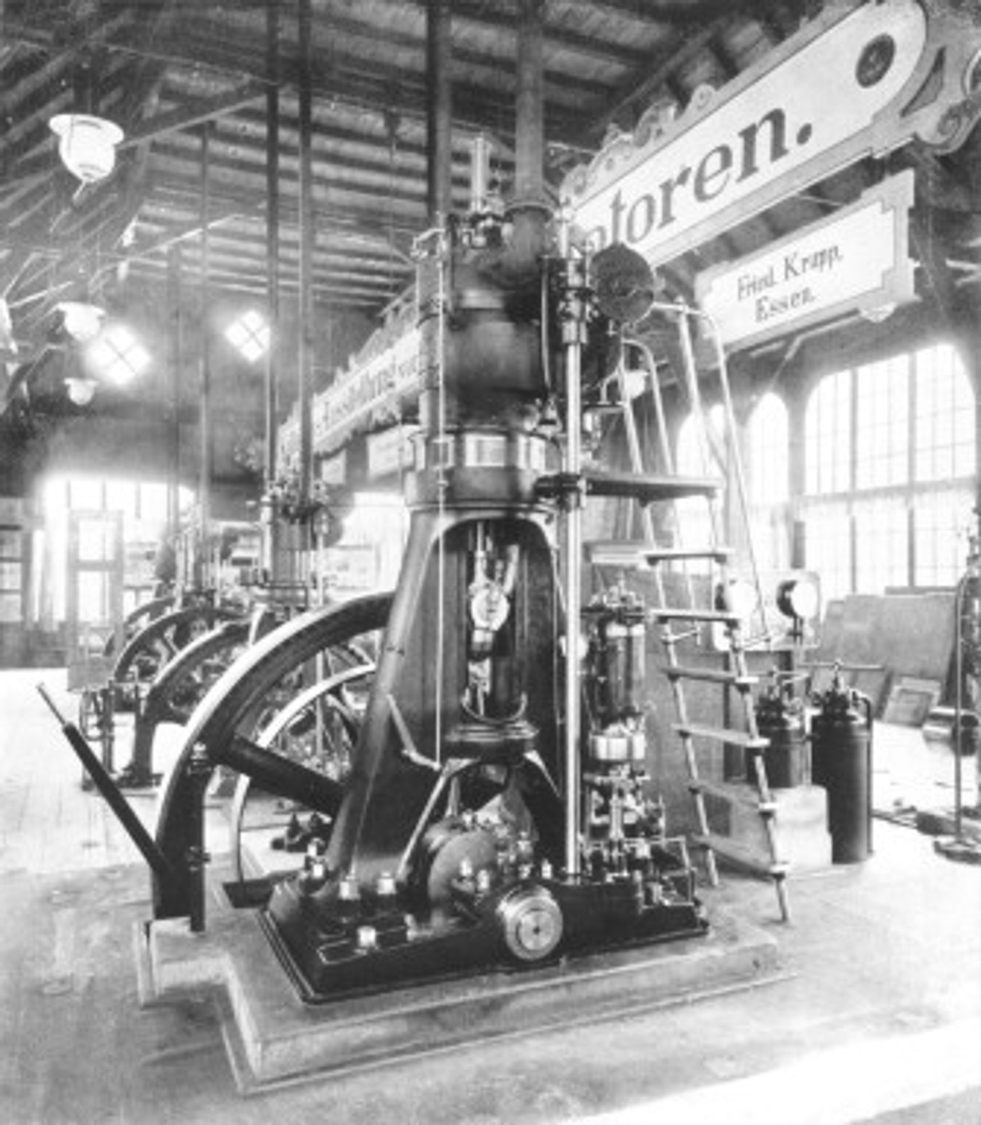 Historische Schwarz-Weiß-Fotografie eines frühen Krupp-Dieselmotors in einer Ausstellungshalle, mit großem Schwungrad und einem Schild mit der Aufschrift „Fried. Krupp, Essen" im Hintergrund, 1898.