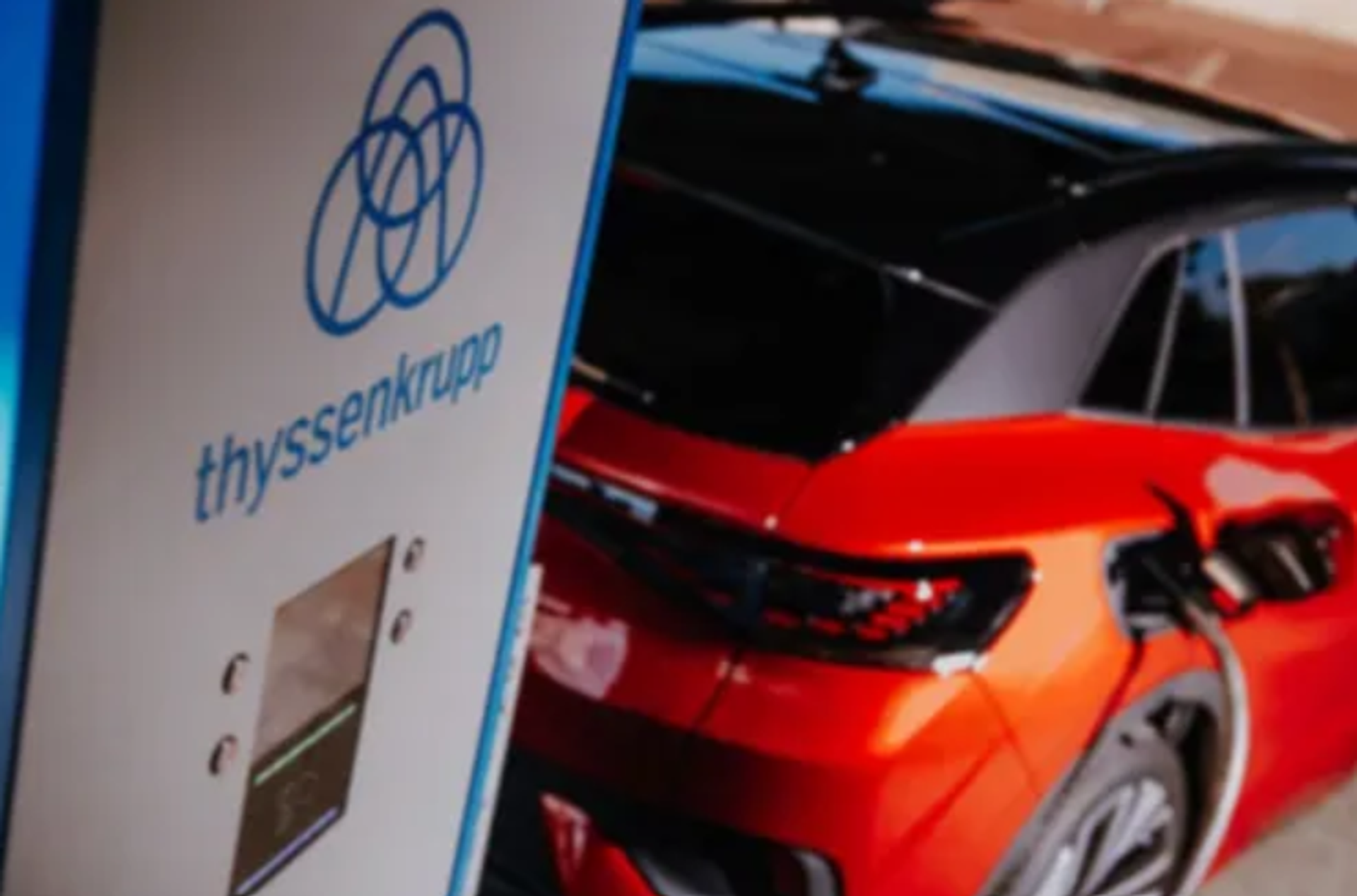 A red electric vehicle is charging at a charging station bearing the thyssenkrupp logo; the charging station is in the foreground